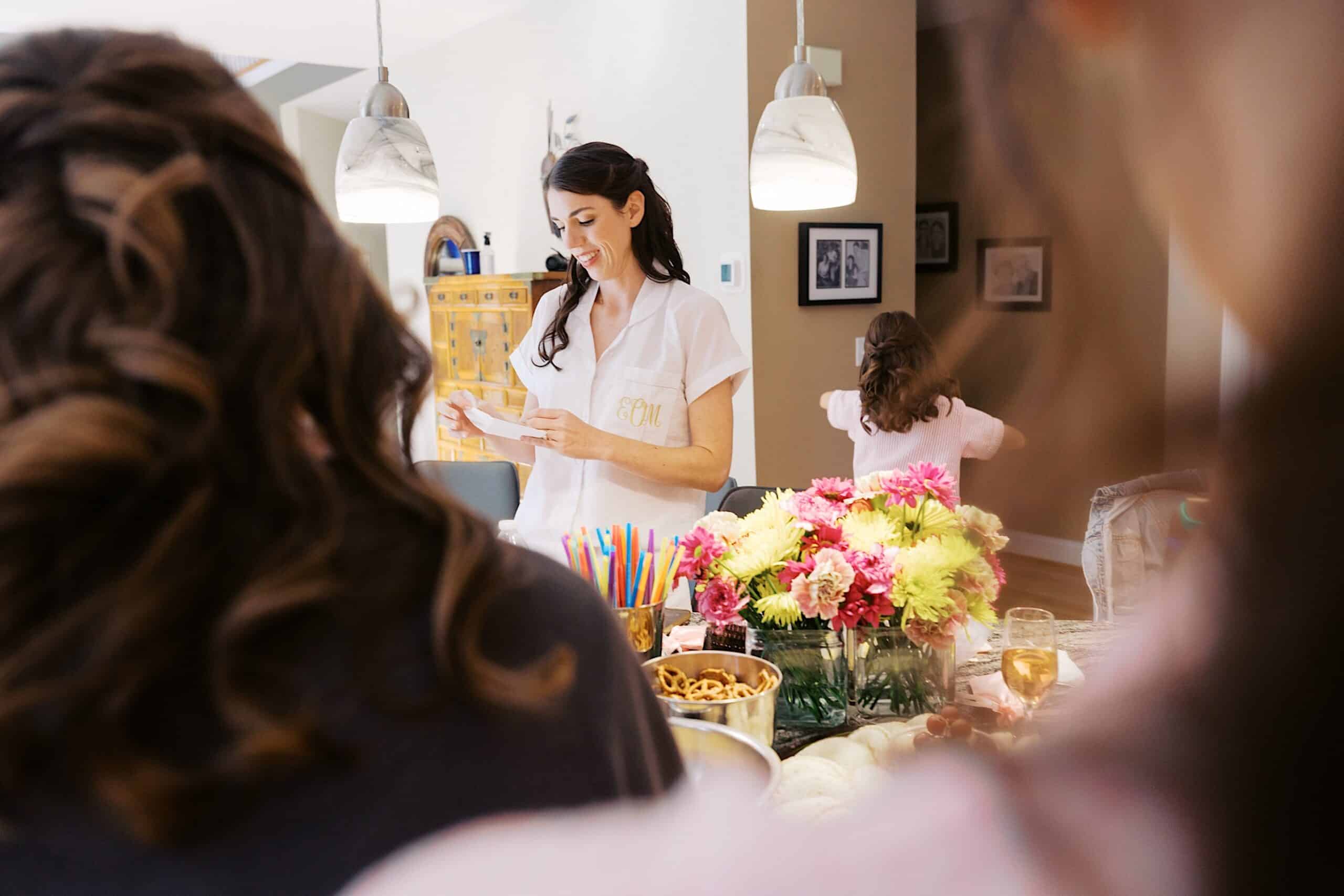 At a luxurious wedding at The Ivy at Ellis Preserve, a woman stands at a decorated table reading from a card, surrounded by flowers, snacks, and drinks. Two people are in the foreground and a child is in the background.