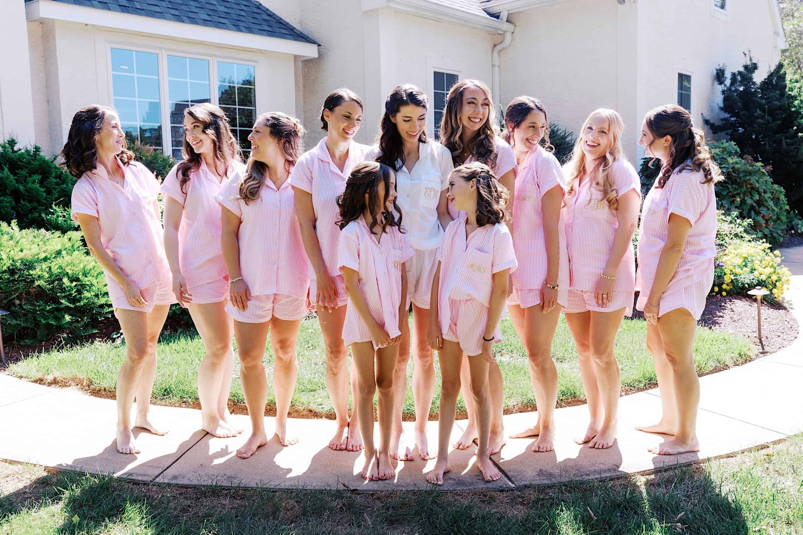 A group of women and girls in matching pink pajamas stand barefoot on a sidewalk outside a house, smiling and chatting—perhaps getting ready for a luxurious wedding at The Ivy at Ellis Preserve.