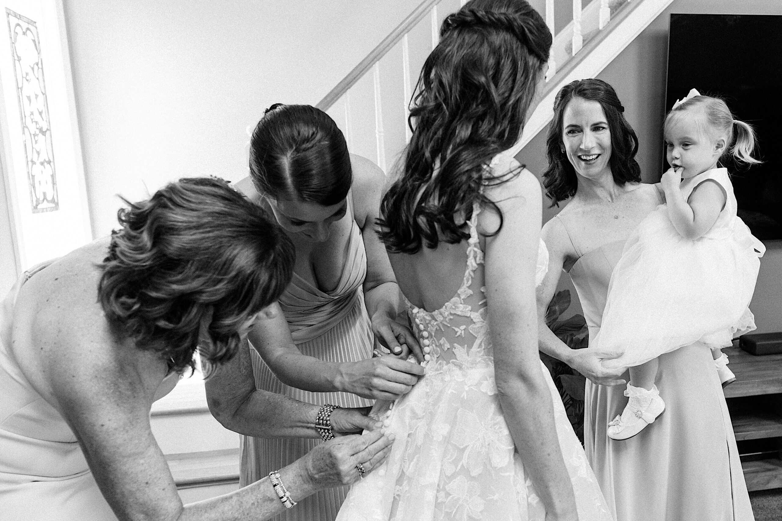 A bride stands as two women fasten her dress while another woman holding a young girl in a dress looks on, capturing an intimate moment before a luxurious wedding at The Ivy at Ellis Preserve.