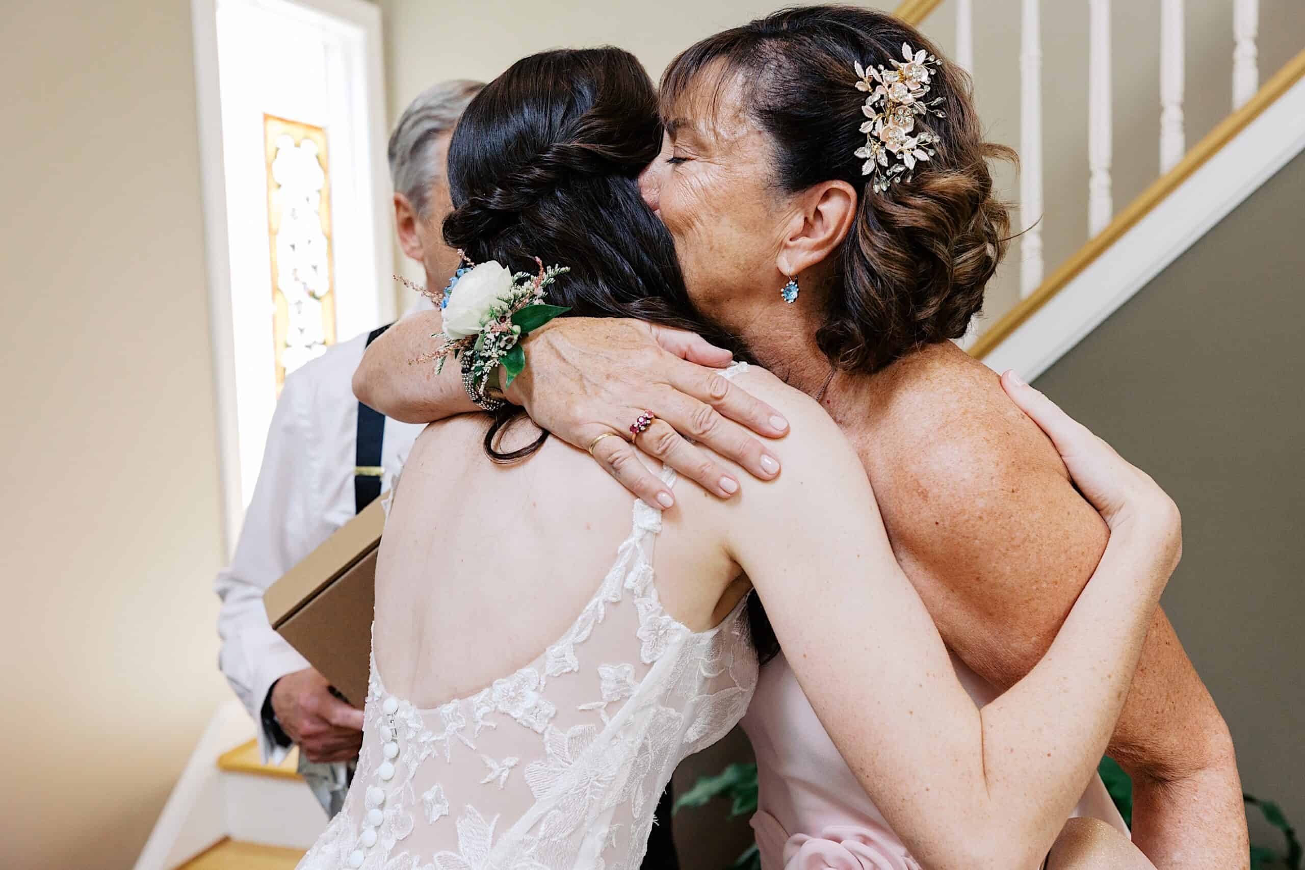 A woman in a wedding dress hugs another woman wearing a floral hairpiece near a staircase indoors during a luxurious wedding at The Ivy at Ellis Preserve.