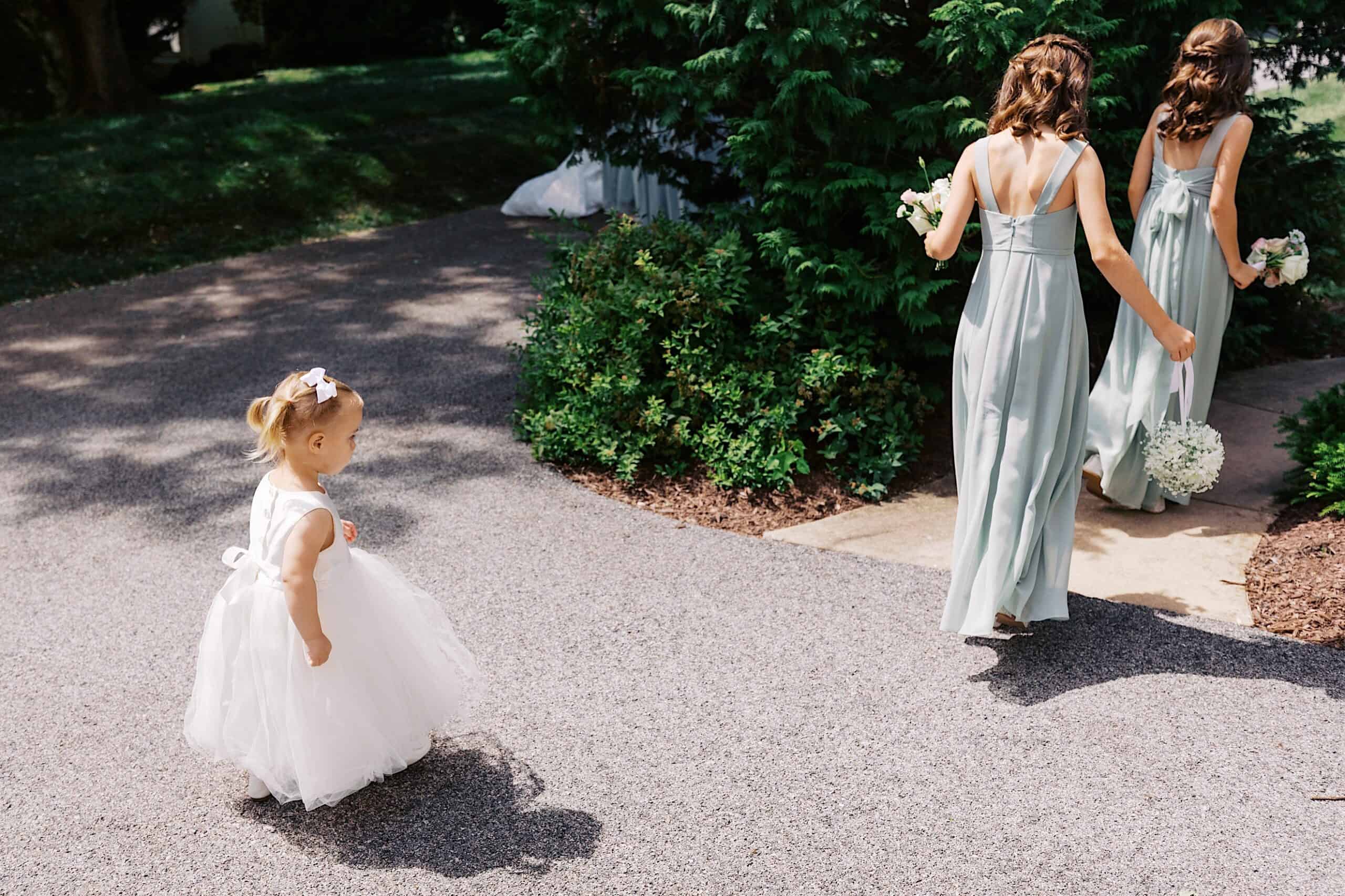A young girl in a white dress walks on a paved path, following two women in light green dresses holding bouquets at a luxurious wedding at The Ivy at Ellis Preserve.