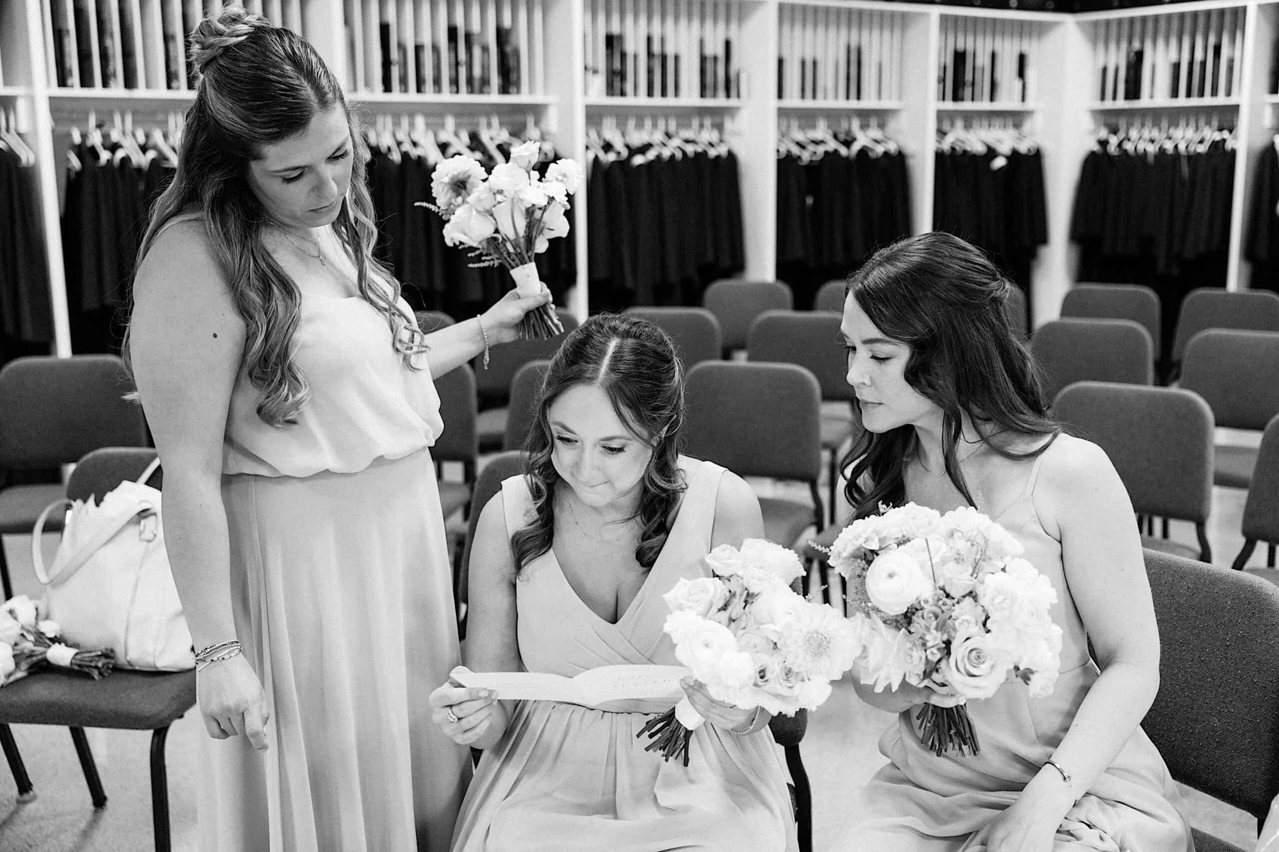 Three women in dresses, two holding bouquets, sit and stand in a room lined with suits and empty chairs during a luxurious wedding at The Ivy at Ellis Preserve. One woman is reading from an open book.