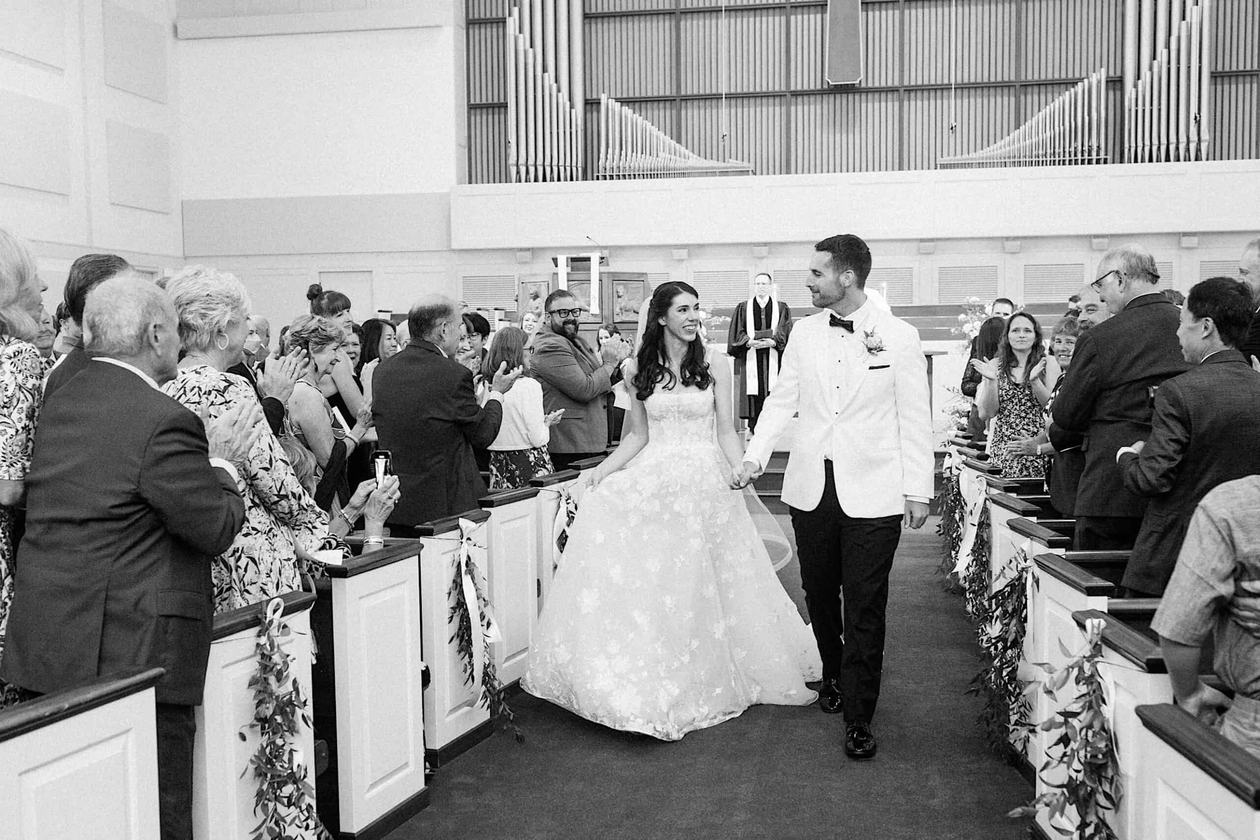 A bride and groom walk down the aisle in a church, holding hands and smiling, while guests stand and applaud on both sides during their luxurious wedding at The Ivy at Ellis Preserve.
