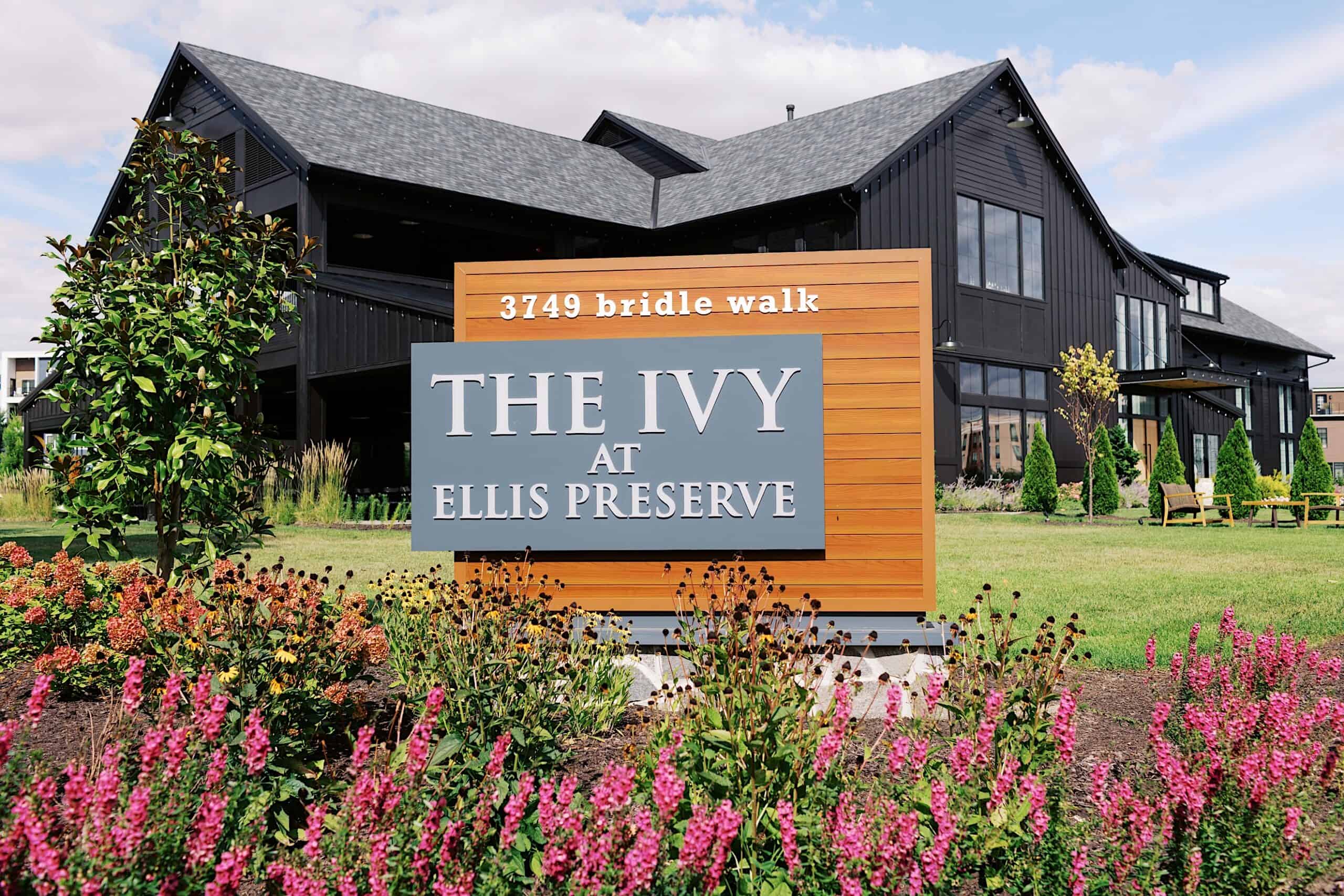 A large black building with many windows stands behind a sign that reads "THE IVY AT ELLIS PRESERVE, 3749 bridle walk," surrounded by landscaping and flowers—the perfect setting for a luxurious wedding at The Ivy at Ellis Preserve.