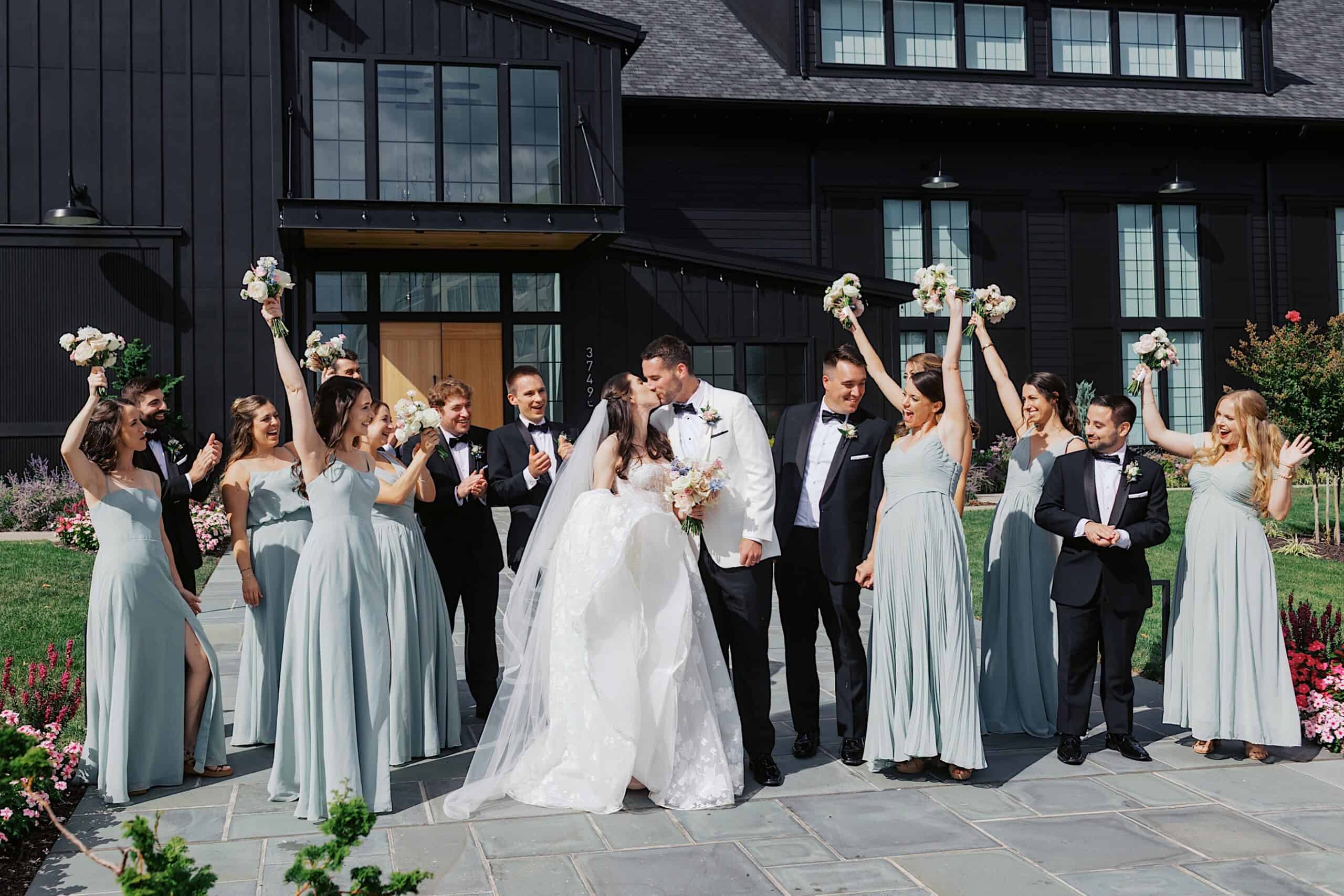 A wedding party poses outdoors at a luxurious wedding at The Ivy at Ellis Preserve; the bride and groom stand in the center, surrounded by bridesmaids in light blue dresses and groomsmen in black suits, some raising bouquets.