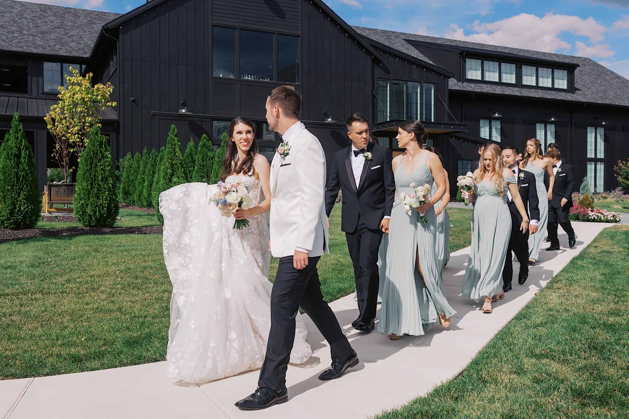 A bride and groom walk on a sidewalk outside a modern black building, accompanied by their wedding party in formal attire during their luxurious wedding at The Ivy at Ellis Preserve.