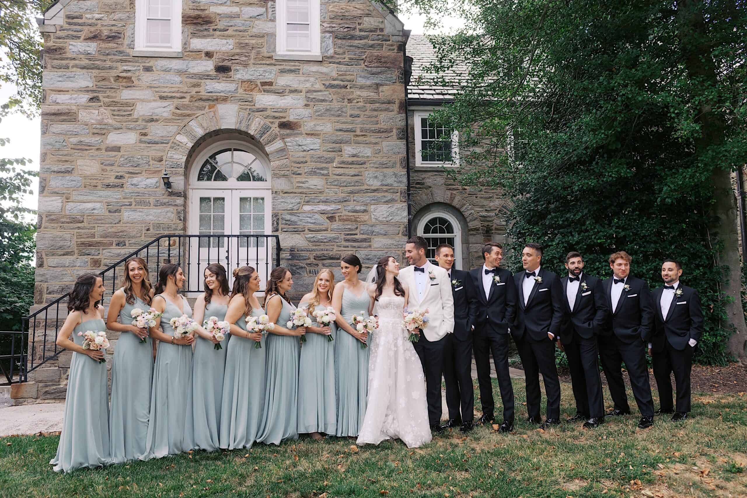 A luxurious wedding at The Ivy at Ellis Preserve: the wedding party stands outside a stone building, bridesmaids in light blue dresses, groomsmen in black suits, with the bride and groom elegantly posed at the center.