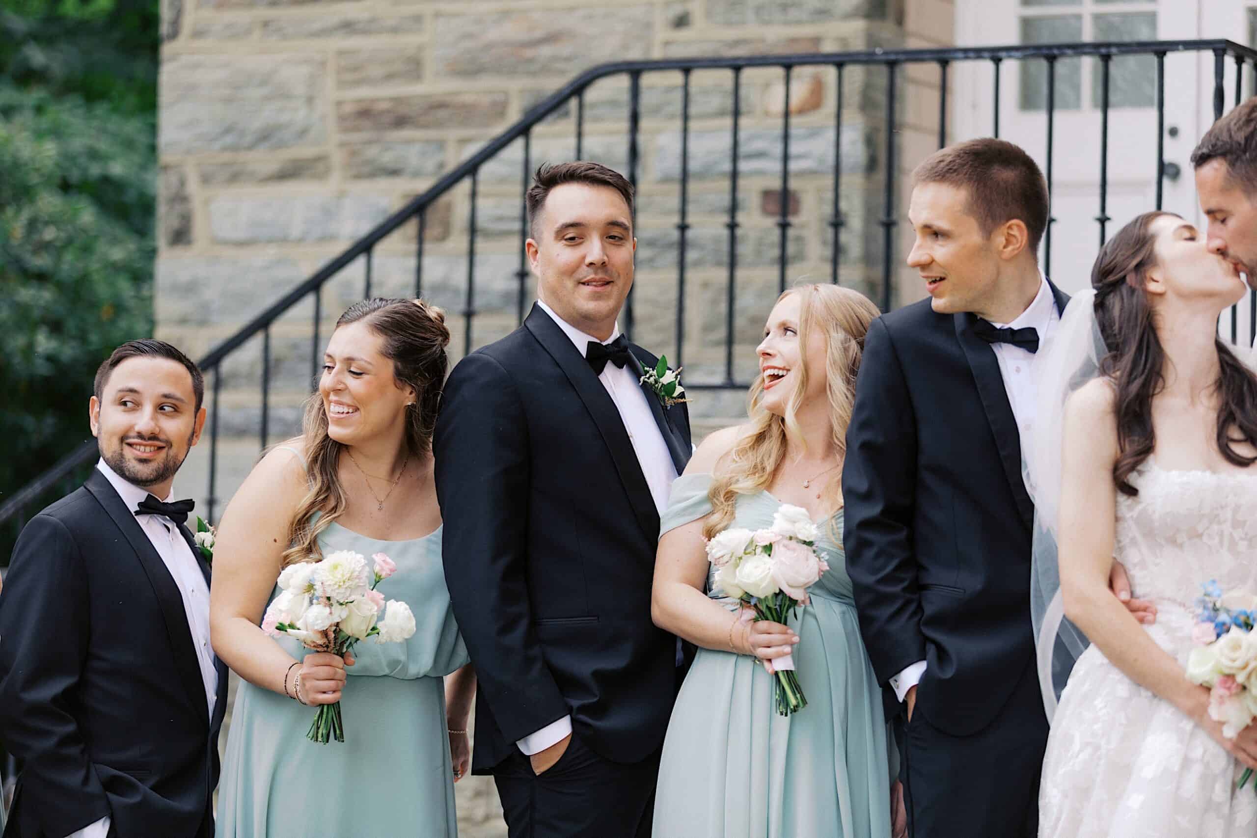 A wedding party poses outdoors at a luxurious wedding at The Ivy at Ellis Preserve; three men in black tuxedos and three women in light green dresses hold bouquets and smile in front of a stone wall with a staircase.