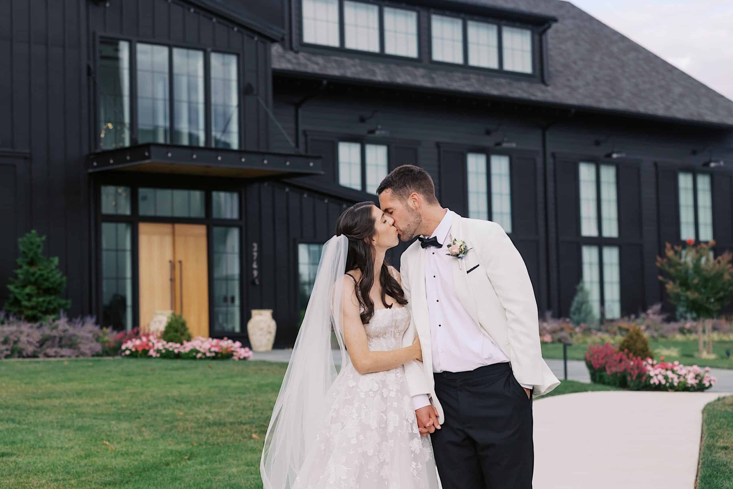 A couple dressed in wedding attire shares a kiss outside The Ivy at Ellis Preserve, capturing the essence of a luxurious wedding with its modern black building, large windows, and manicured lawn.