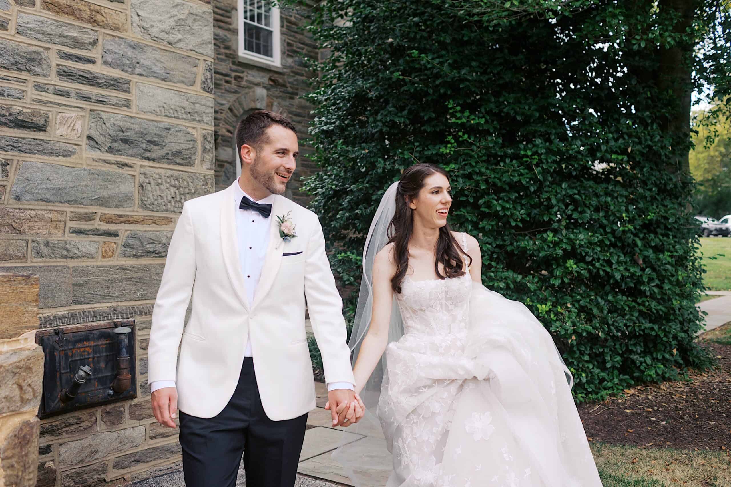 A bride in a strapless white gown and a groom in a white tuxedo jacket hold hands and walk outside near a stone building and greenery, capturing the elegance of their luxurious wedding at The Ivy at Ellis Preserve.