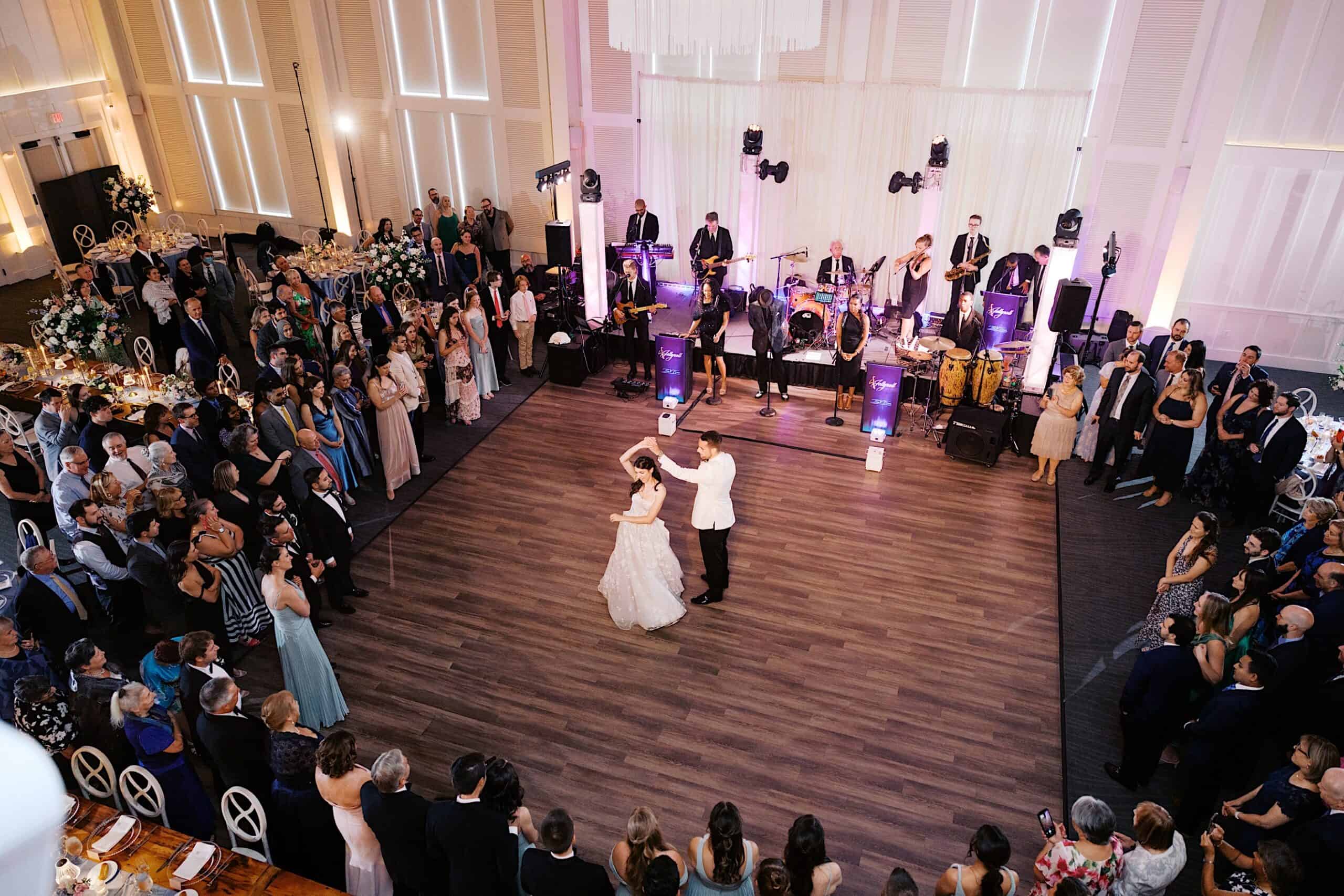 A bride and groom dance in the center of a large ballroom surrounded by guests, with a live band performing on stage in the background at a luxurious wedding at The Ivy at Ellis Preserve.