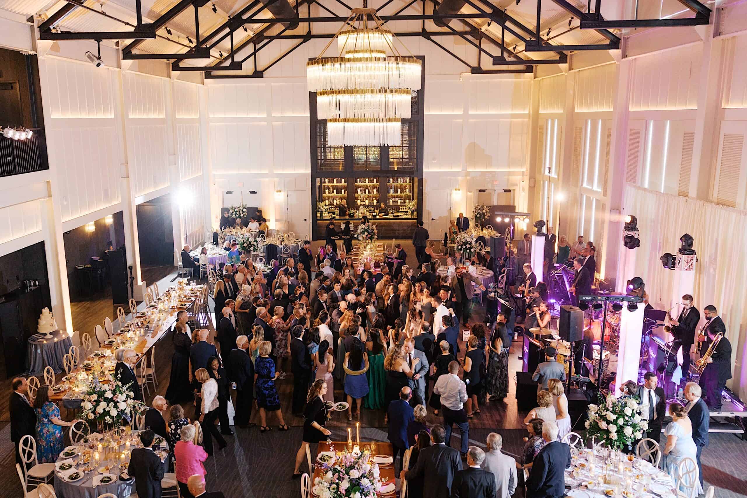 A large group attends a luxurious wedding at The Ivy at Ellis Preserve, enjoying a formal event in an elegant ballroom with chandeliers, beautifully decorated tables, and a live band performing on stage.