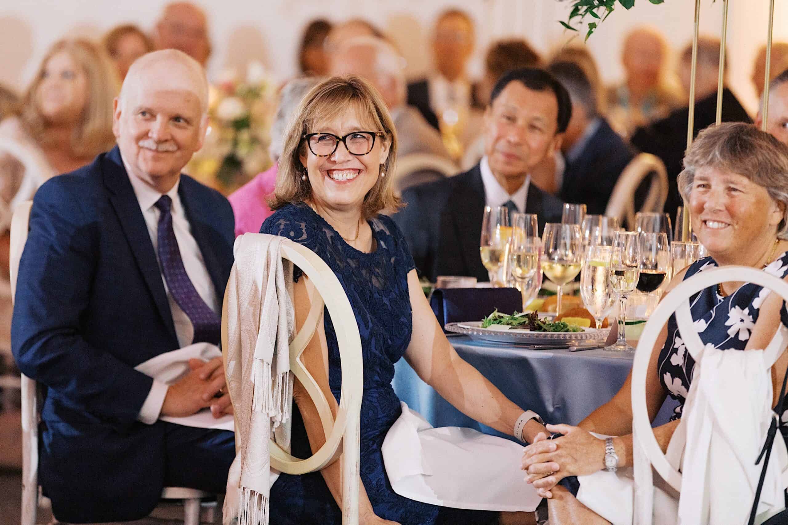 A group of people in formal attire sit at a round table with plates of food and glasses of wine, smiling at the camera during a luxurious wedding at The Ivy at Ellis Preserve.