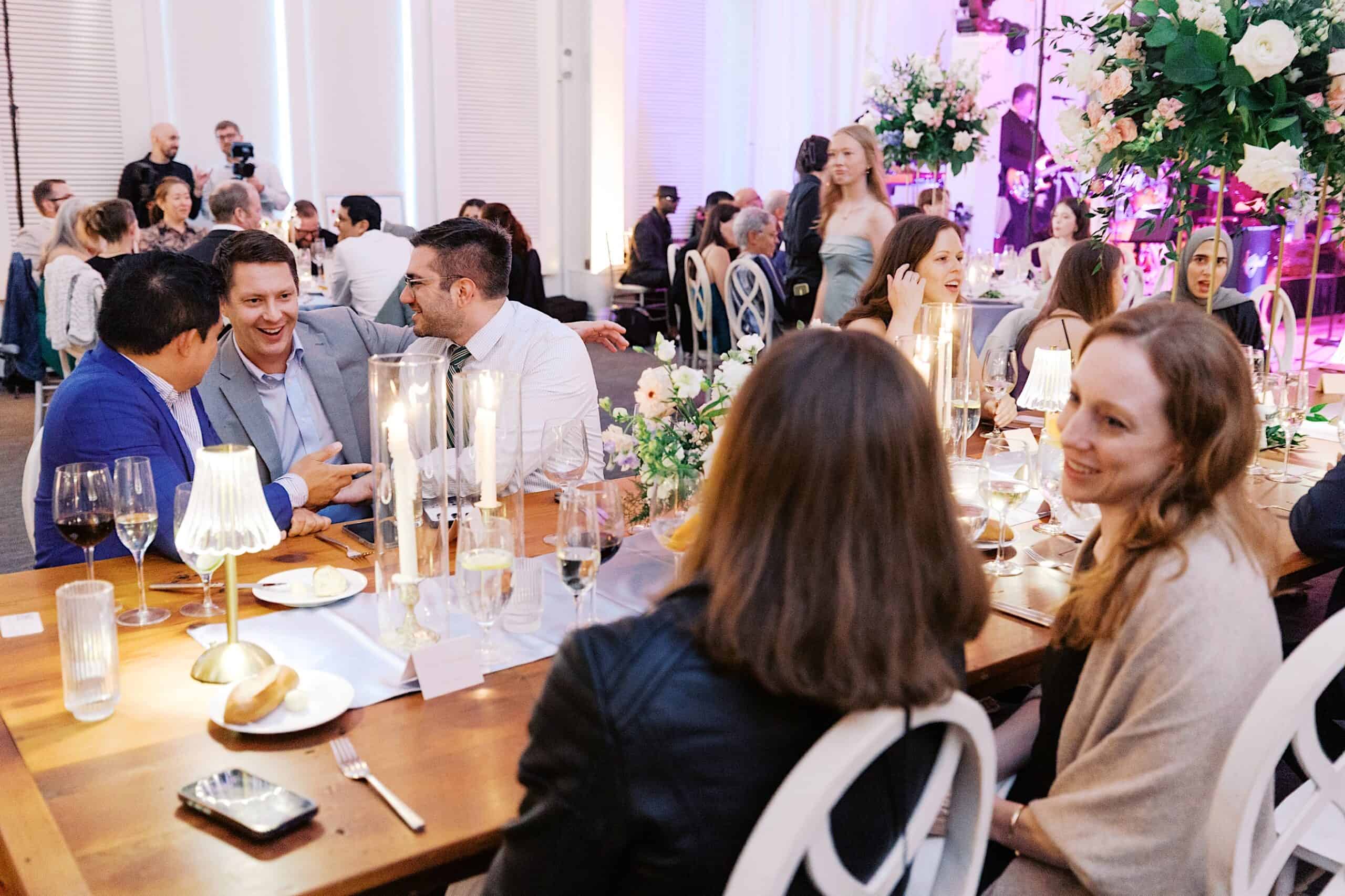 Guests sit at a decorated table with candles and flowers, engaged in conversation during a luxurious wedding at The Ivy at Ellis Preserve.