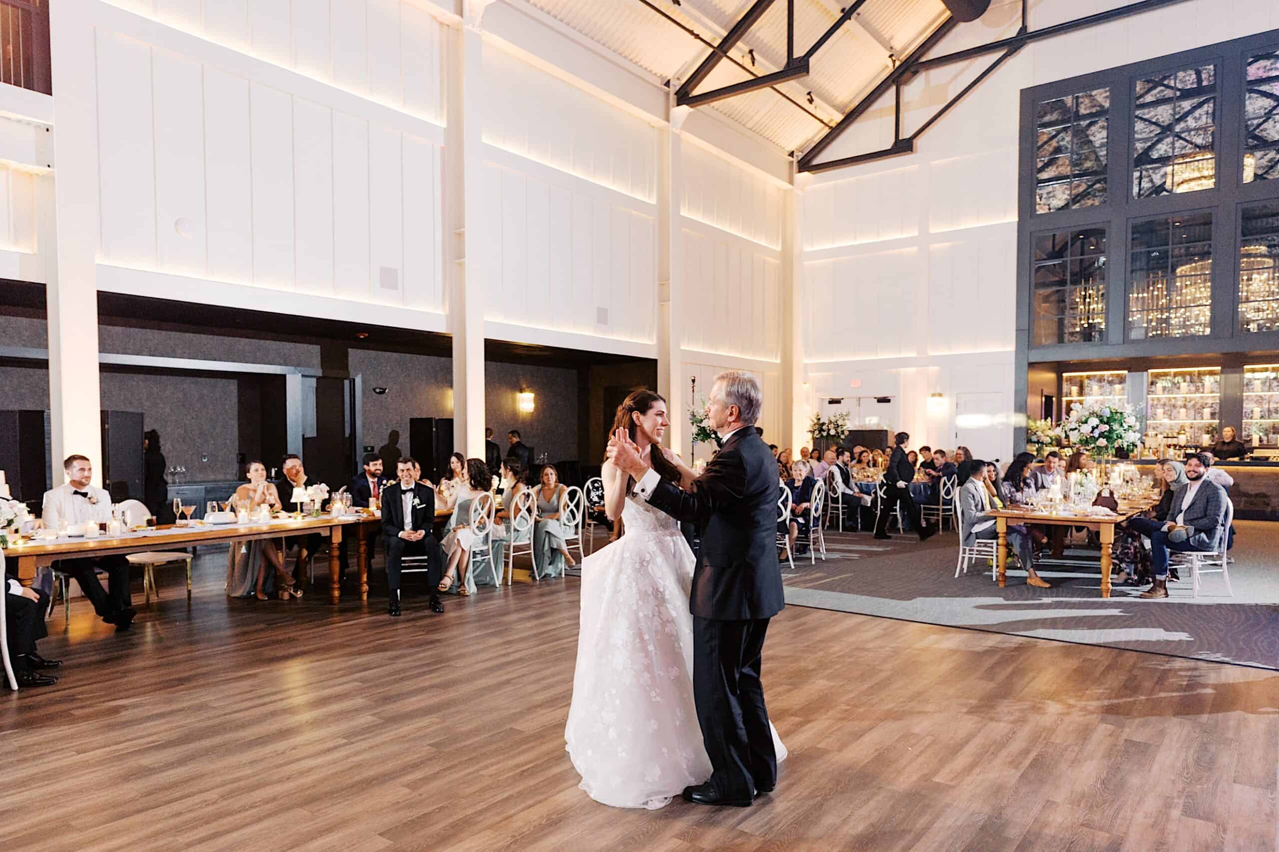 A bride and an older man dance together in the center of a spacious, elegant banquet hall during a luxurious wedding at The Ivy at Ellis Preserve, while guests seated at tables watch.