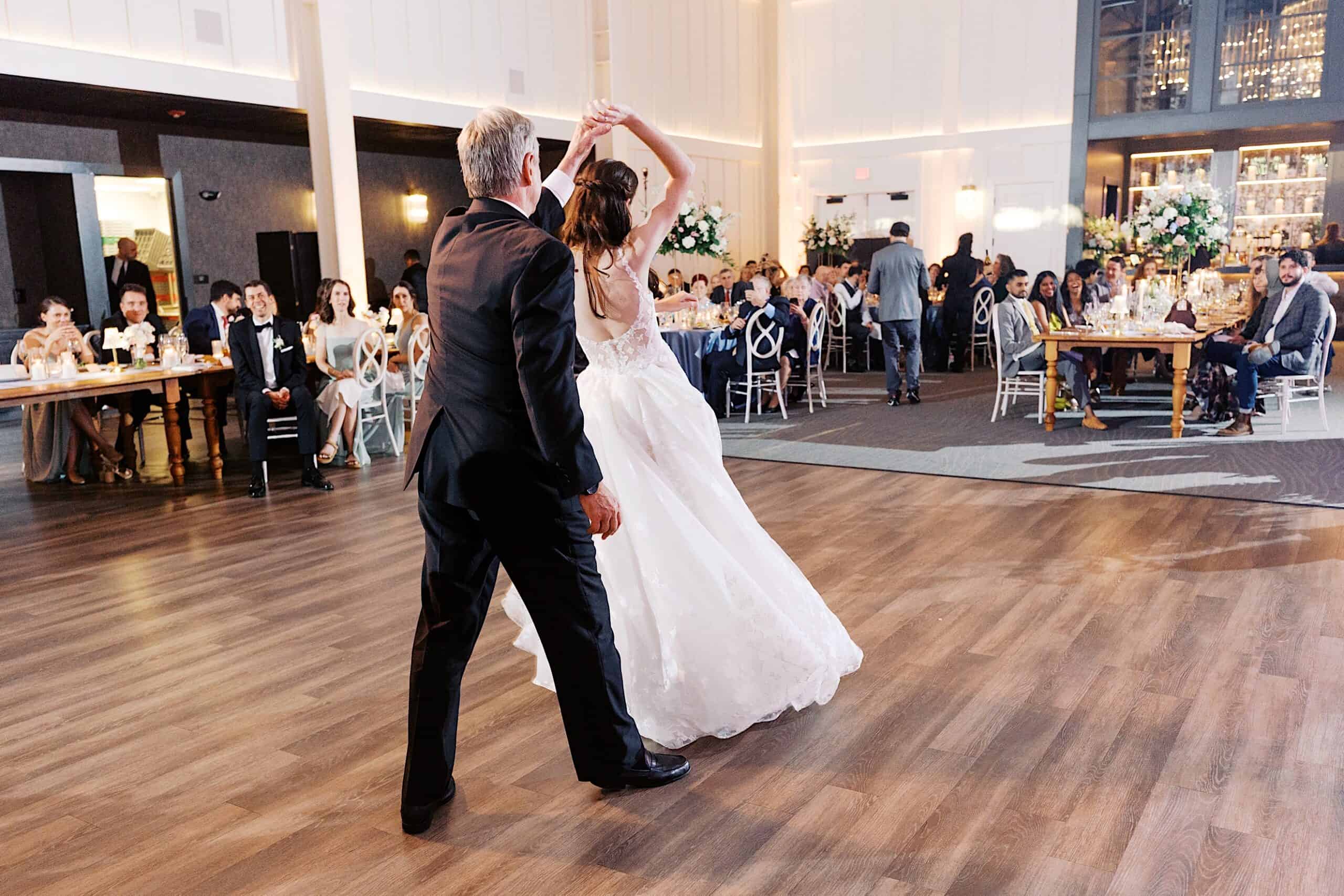A bride in a white gown dances with her father on a wooden floor as guests watch, celebrating a luxurious wedding at The Ivy at Ellis Preserve.