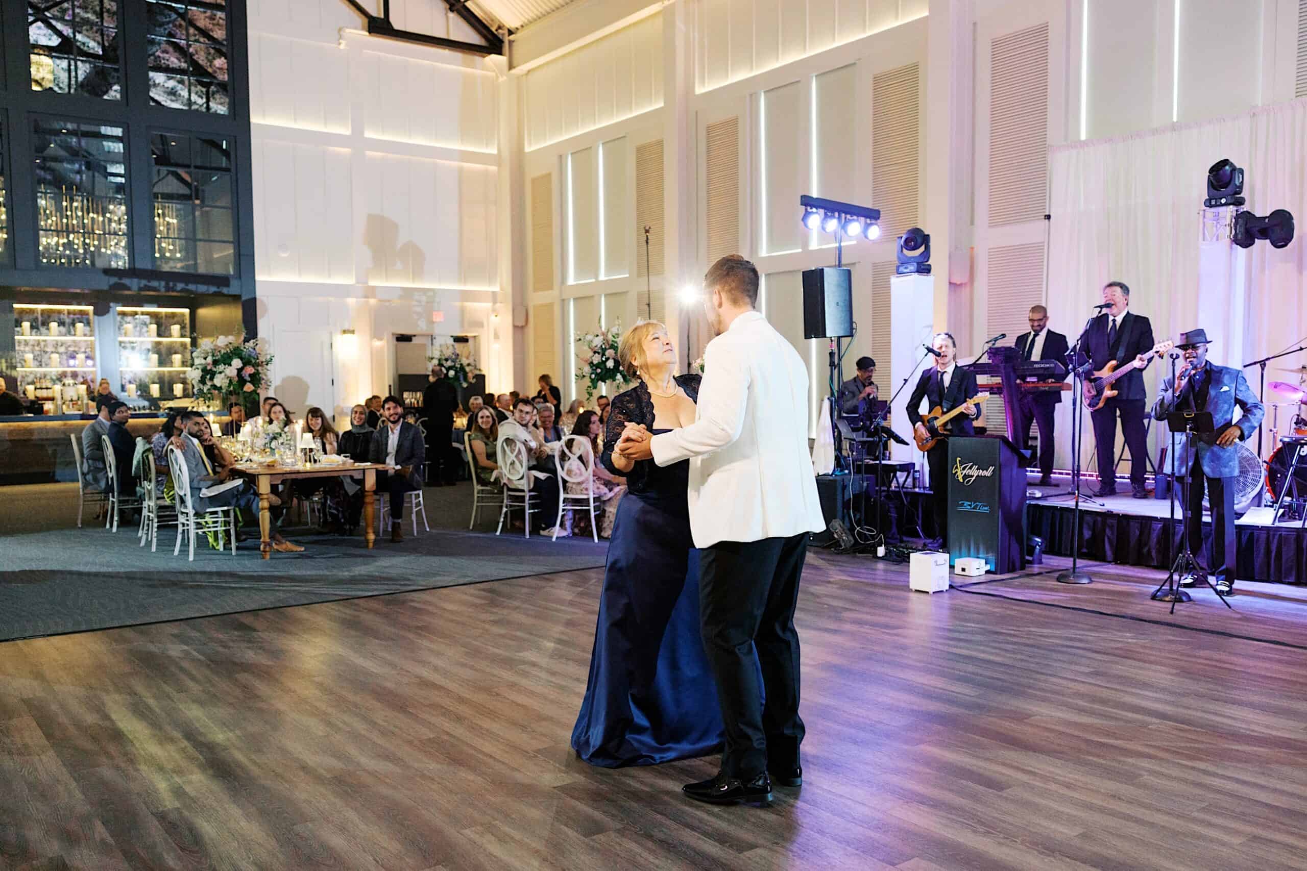 A man in a white jacket dances with an older woman in a navy dress on the floor of a luxurious wedding at The Ivy at Ellis Preserve, while guests enjoy the reception and a live band performs in the background.