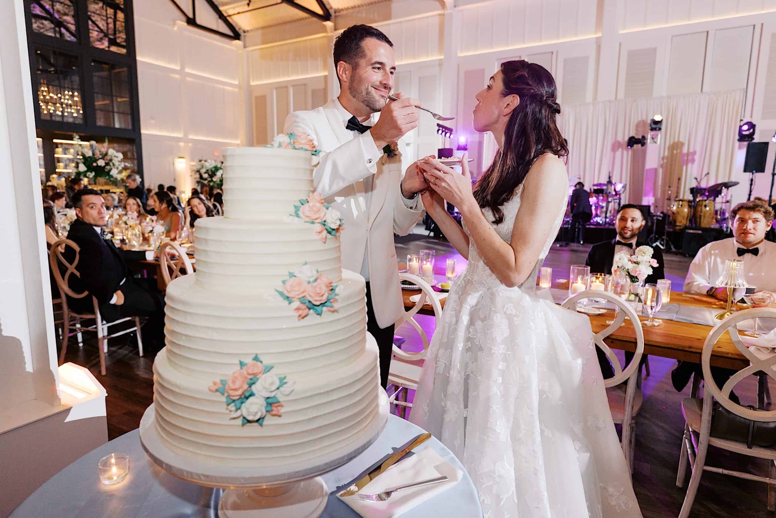 A bride and groom in formal attire feed each other cake at their luxurious wedding at The Ivy at Ellis Preserve, with a large tiered cake in the foreground and guests seated in the background.