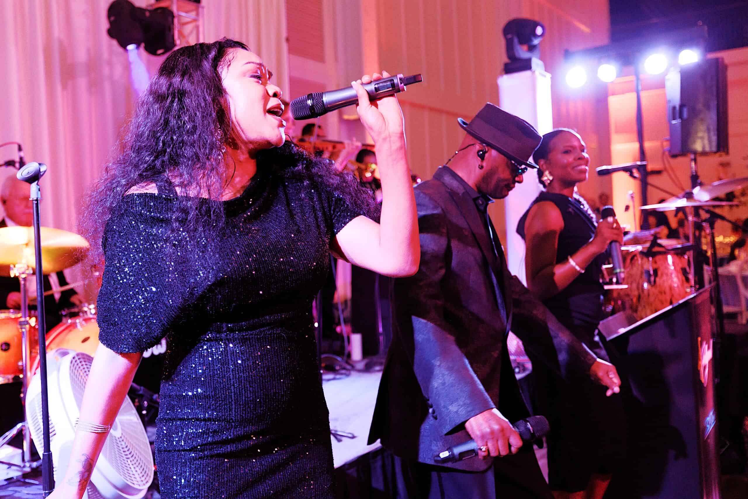 Three musicians perform on stage with microphones under colorful lights at a luxurious wedding at The Ivy at Ellis Preserve; the woman in front wears a sparkly black dress, with the man and second woman standing behind her.