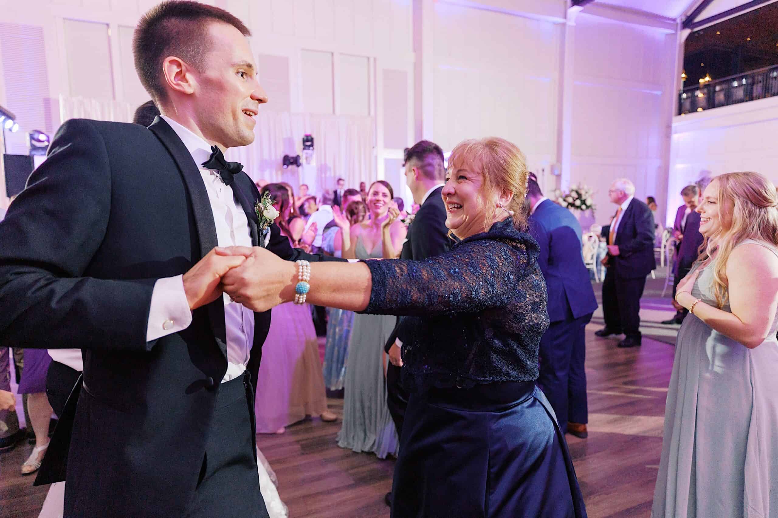 A man in a tuxedo and a woman in a navy dress are holding hands and dancing at a luxurious wedding at The Ivy at Ellis Preserve, with other guests enjoying the elegant indoor celebration in the background.