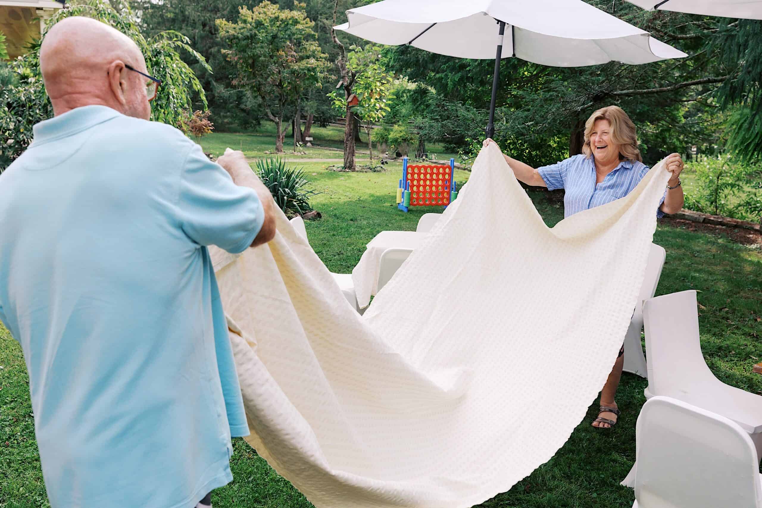 Two adults outdoors hold a large white blanket between them, preparing a seating area with chairs and a table in a green garden for a beautiful wedding at Vincent Forge Mansion.