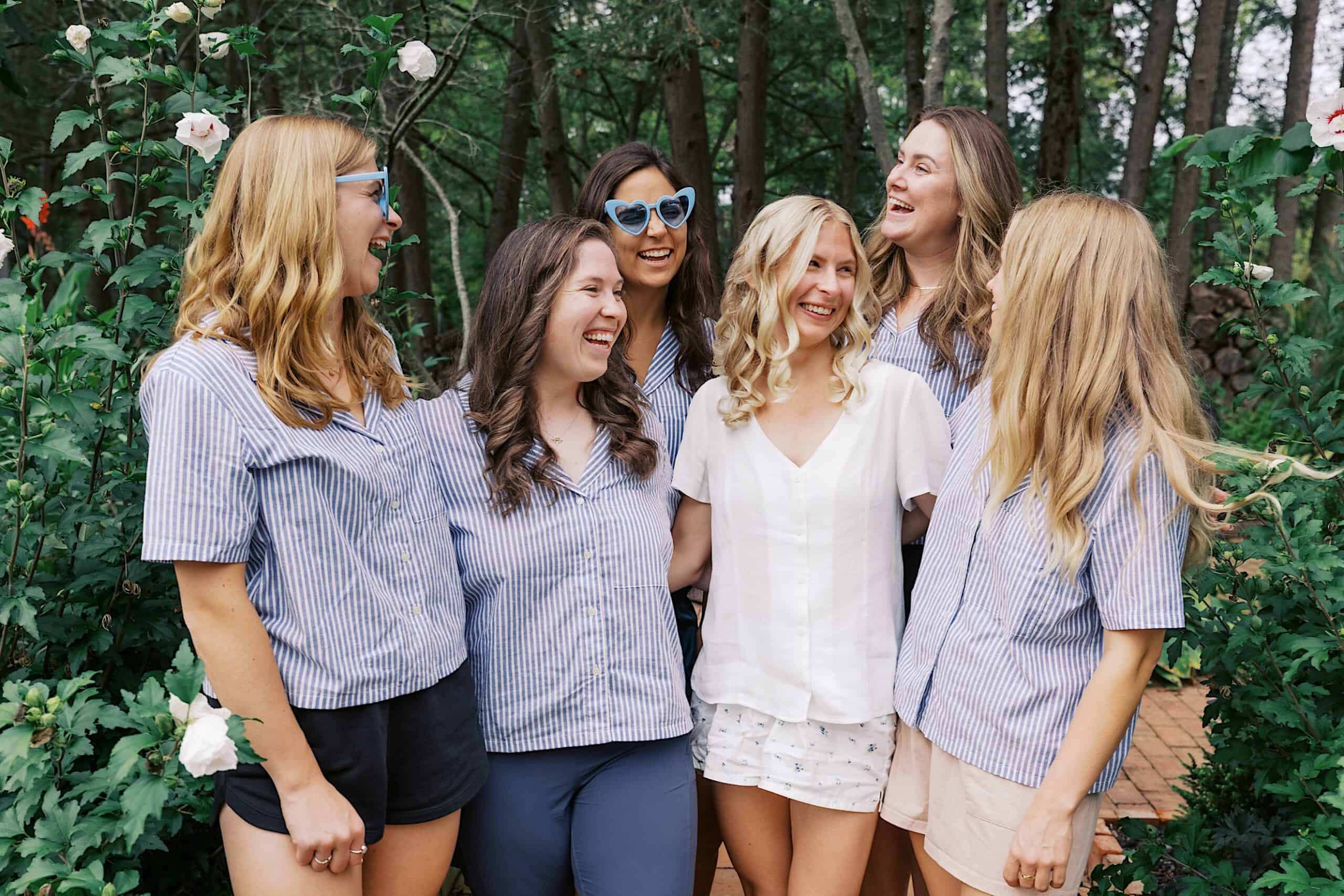 Six women stand together outdoors in a garden at the beautiful wedding at Vincent Forge Mansion, smiling and laughing. Five wear blue striped shirts, one wears a white shirt and shorts. Roses and greenery add charm to the scene.