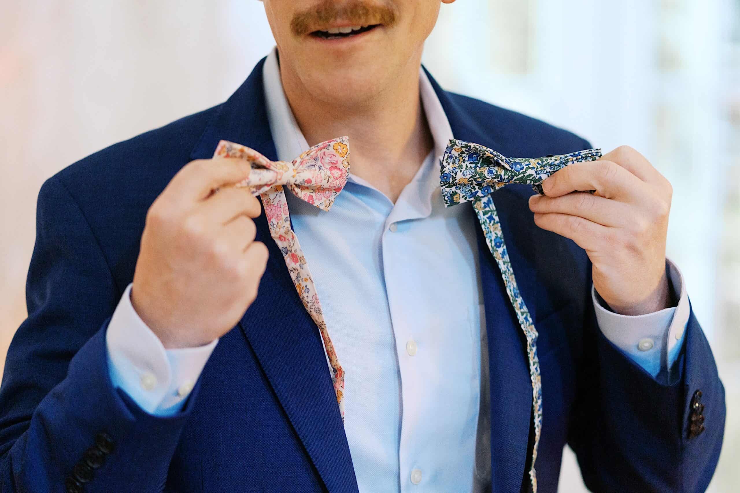 A man in a blue suit holds two patterned bow ties, one pink and one blue, while smiling at a beautiful wedding at Vincent Forge Mansion.