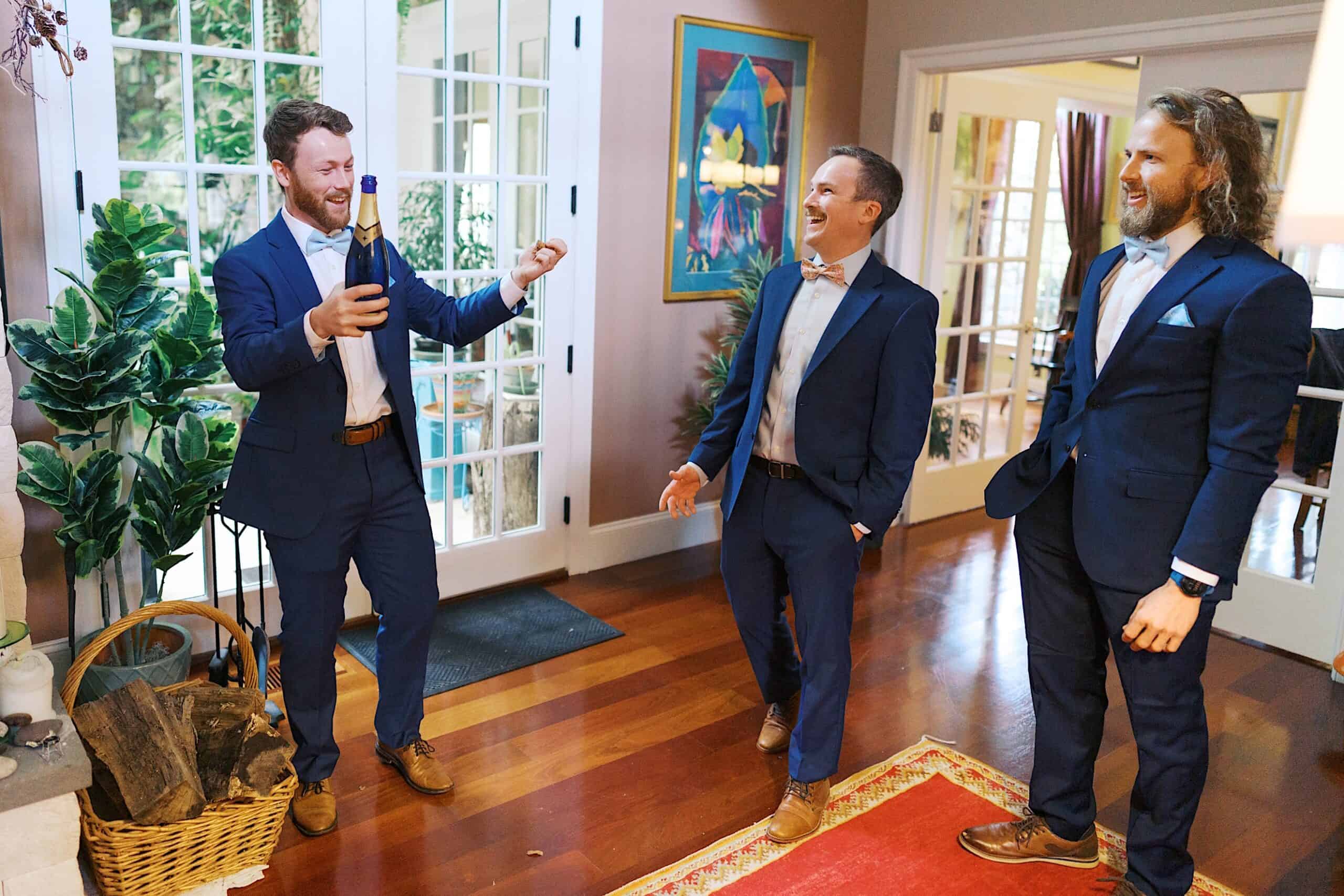 Three men in blue suits stand in a living room during a beautiful wedding at Vincent Forge Mansion, one holding a champagne bottle and smiling as the others watch and laugh.