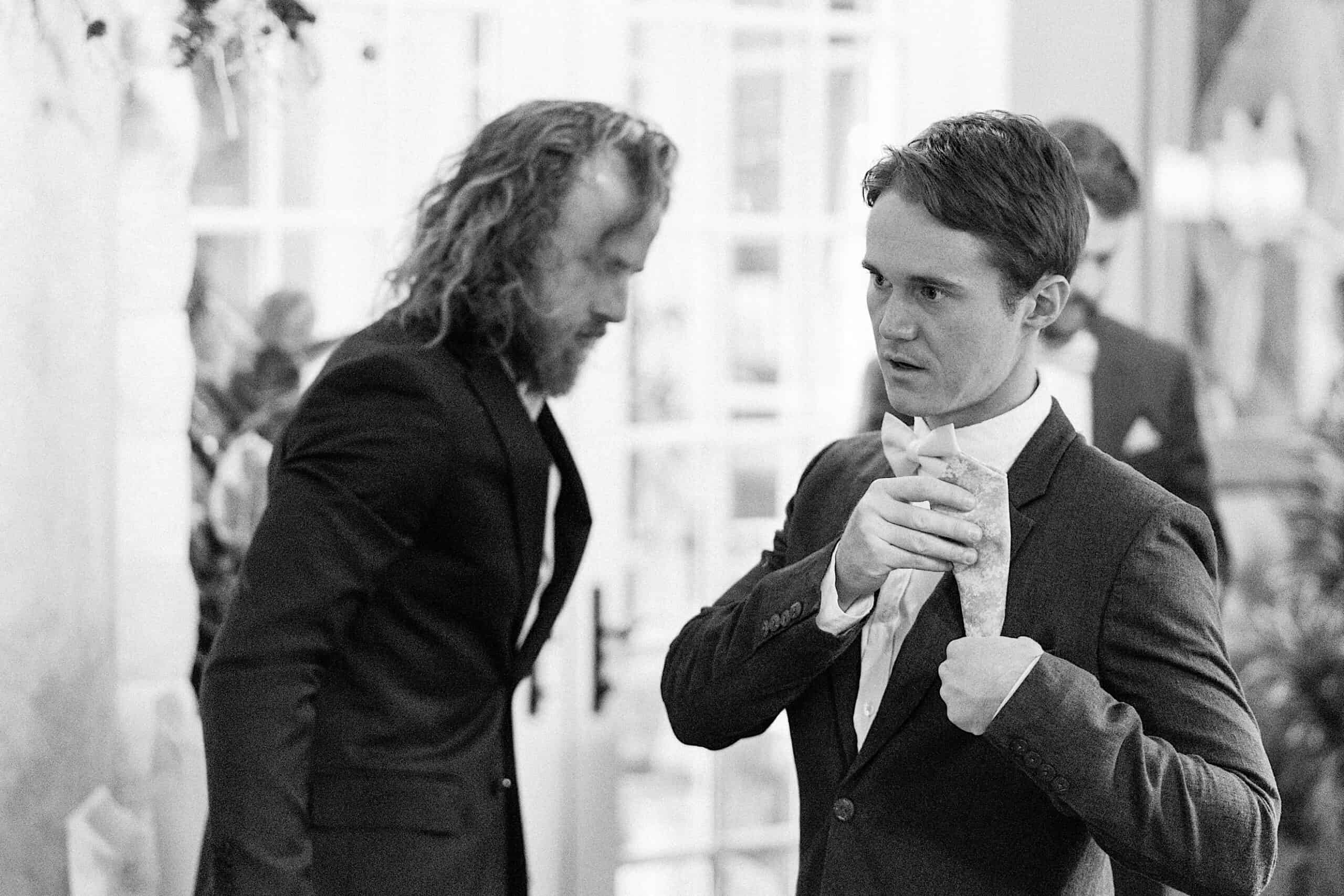 Two men in suits stand indoors at a beautiful wedding at Vincent Forge Mansion; one adjusts his tie while the other looks down, slightly blurred in the background.