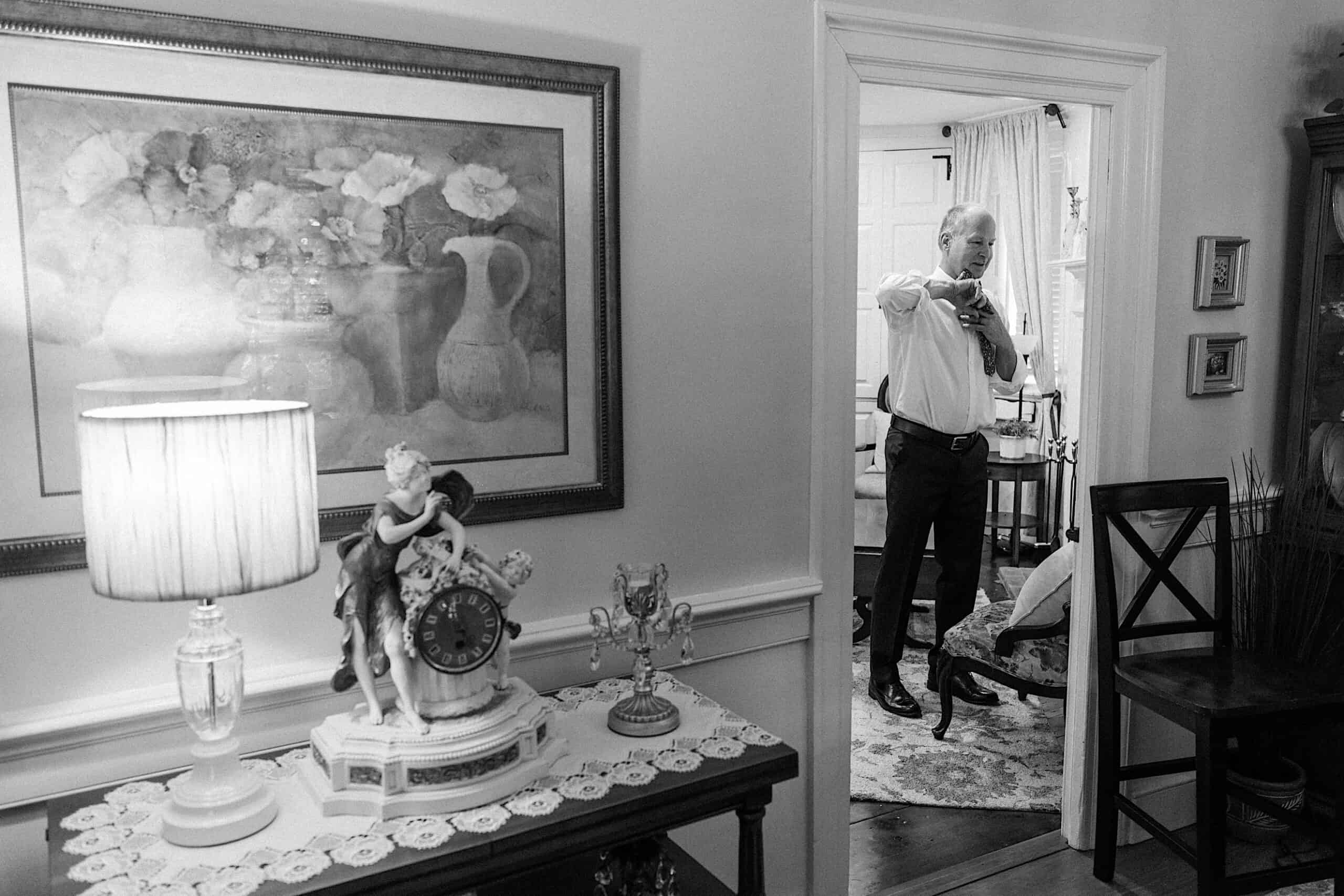 A man adjusts his tie in a room, viewed through a doorway from an adjacent room decorated with a lamp, clock, and framed flower painting, preparing for a beautiful wedding at Vincent Forge Mansion.