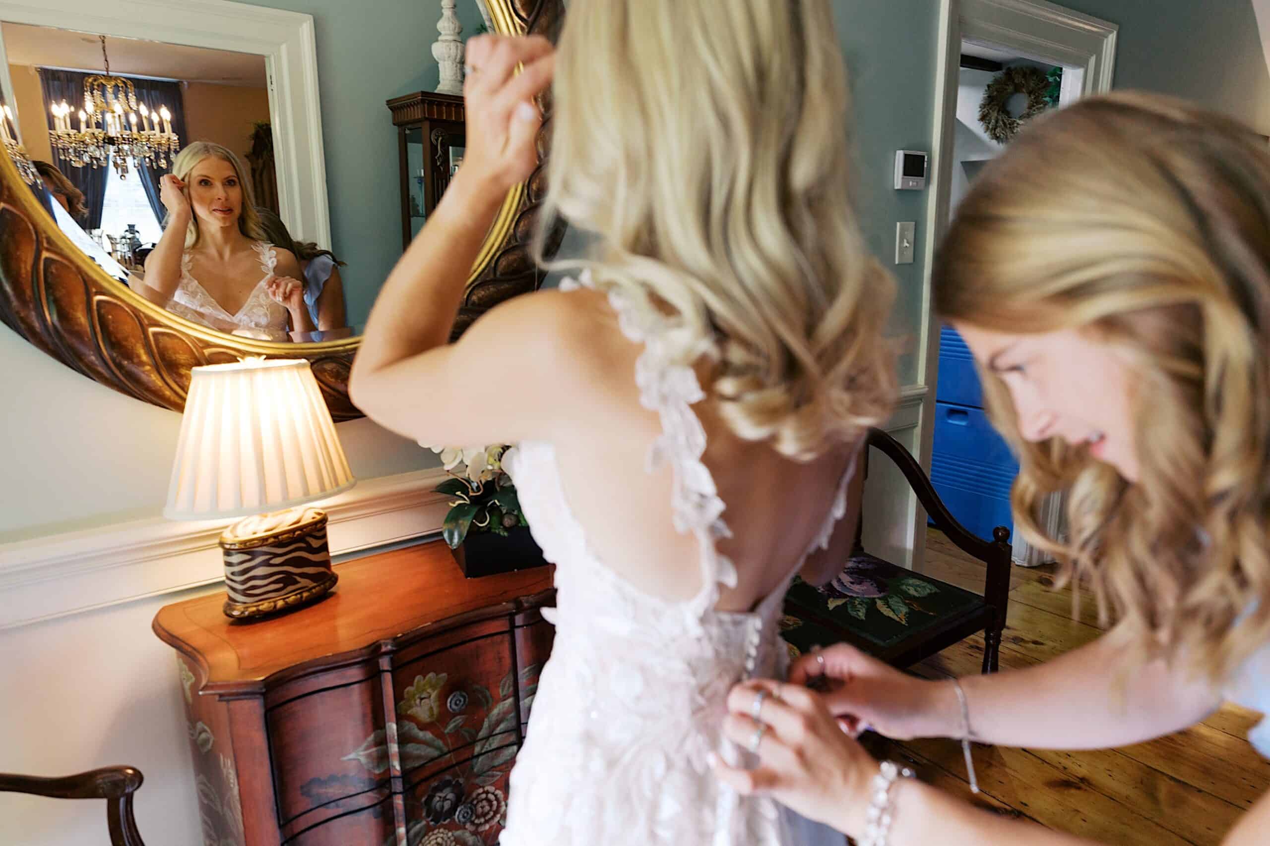 A woman fastens the back of a bride’s dress as the bride admires her reflection in a well-lit room, preparing for a beautiful wedding at Vincent Forge Mansion.