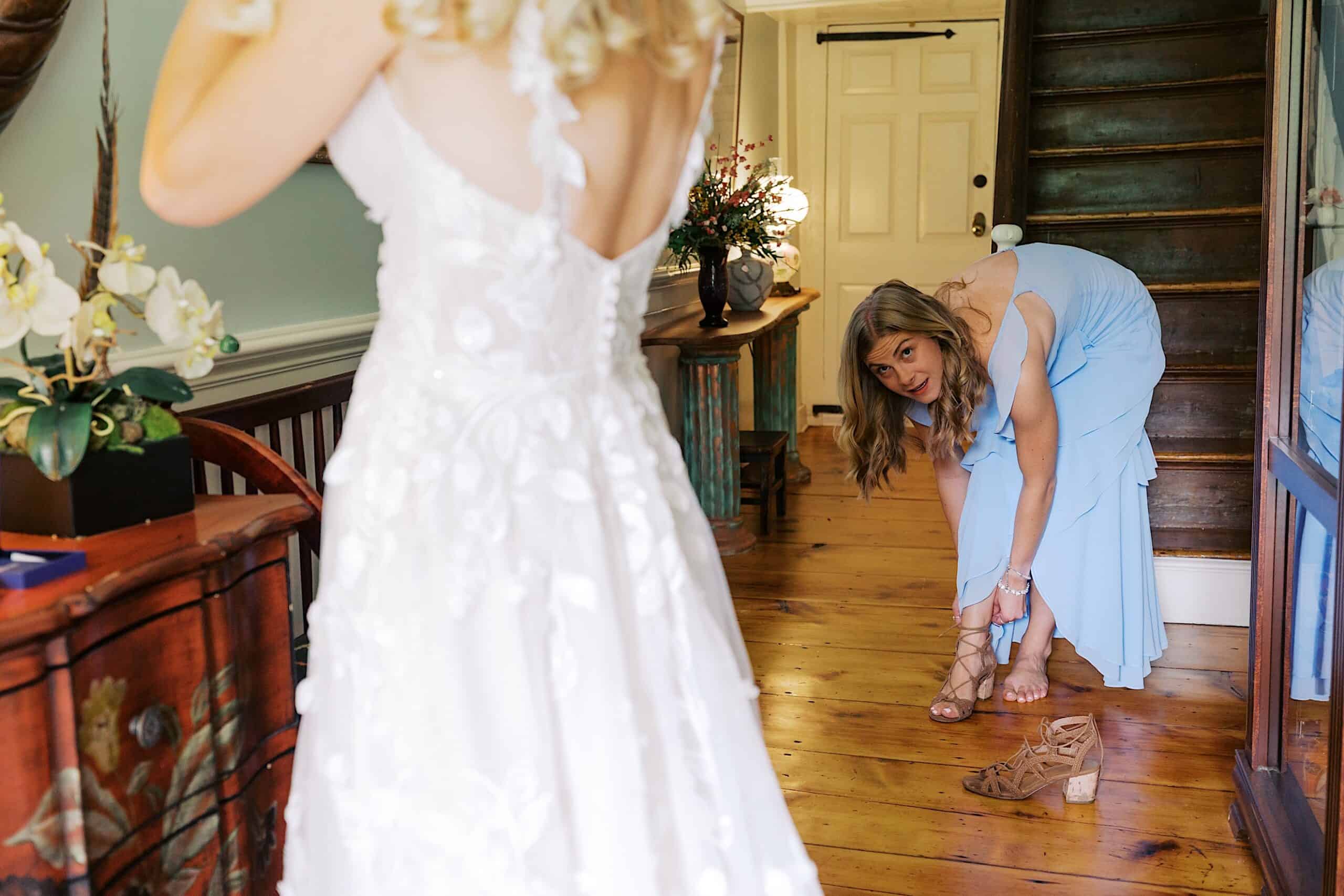 A woman in a blue dress bends down to fasten her sandal while another woman in a white dress stands nearby in a hallway with wooden floors at a beautiful wedding at Vincent Forge Mansion.