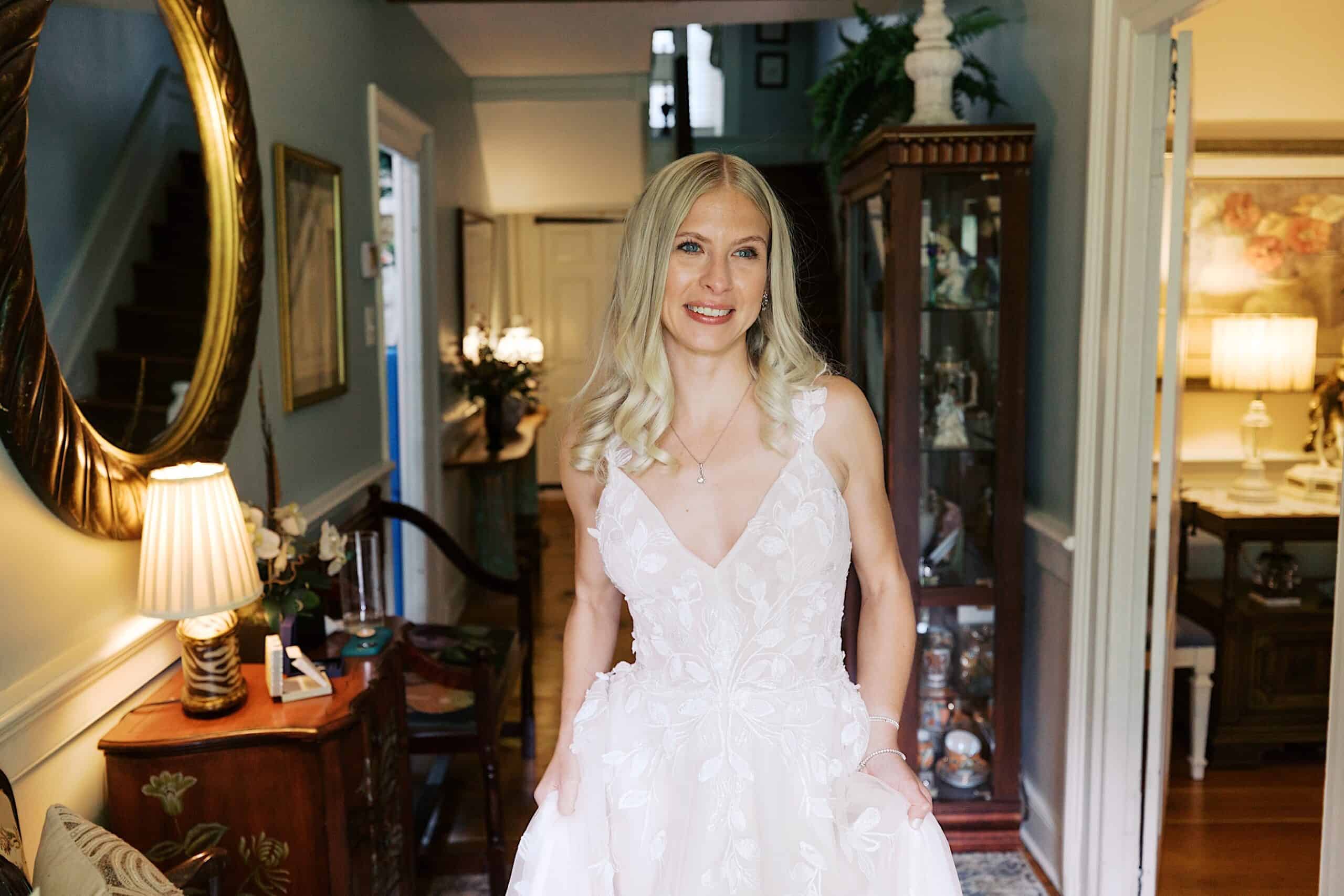 A woman in a white, floral-patterned dress stands in a well-lit, traditionally decorated living room at the beautiful wedding at Vincent Forge Mansion, smiling at the camera.