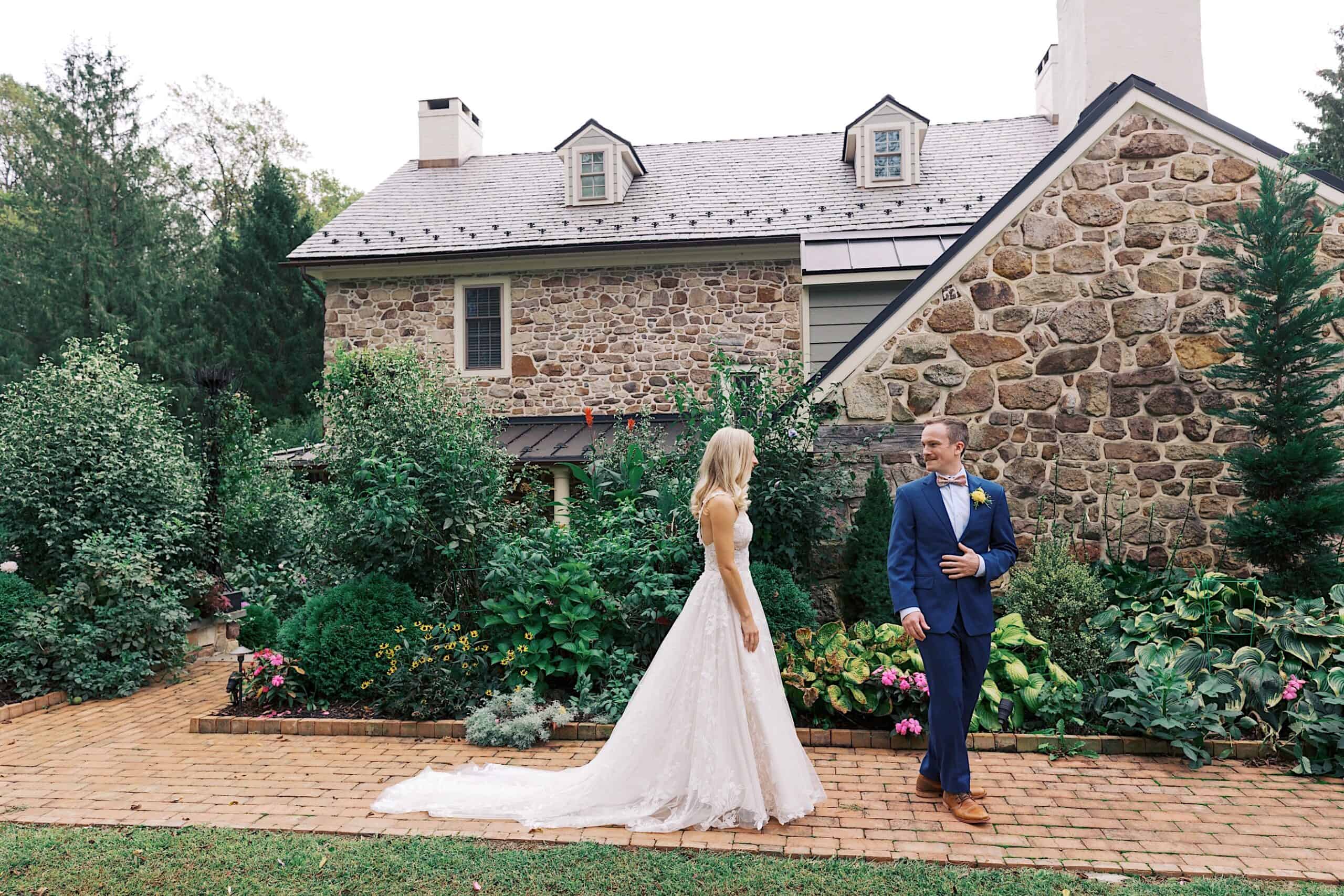A bride and groom stand facing each other on a brick path in front of the stone house at Vincent Forge Mansion, surrounded by green plants and flowers, making for a beautiful wedding setting.