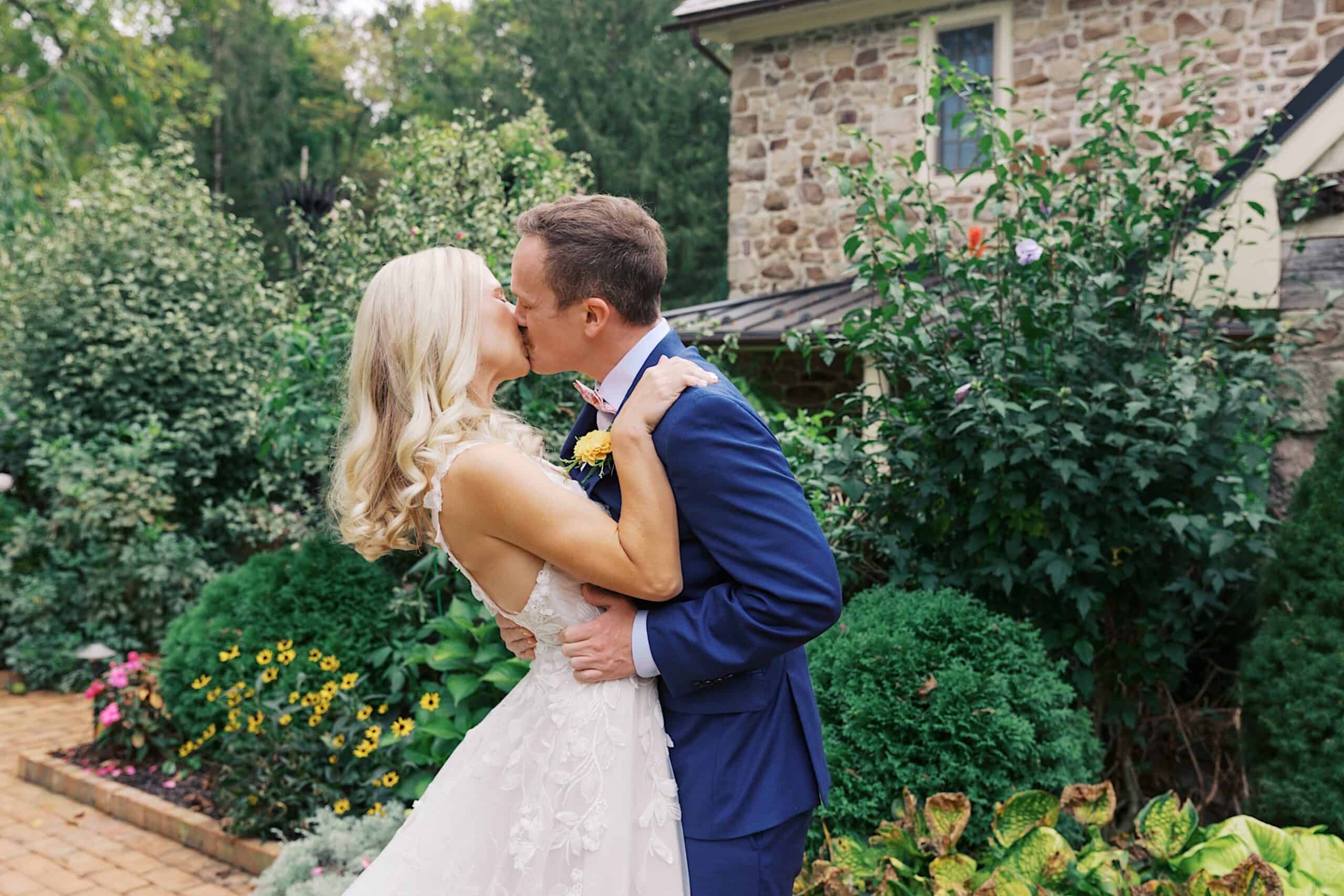 A bride and groom kiss outdoors in a garden setting in front of a stone house during their beautiful wedding at Vincent Forge Mansion.