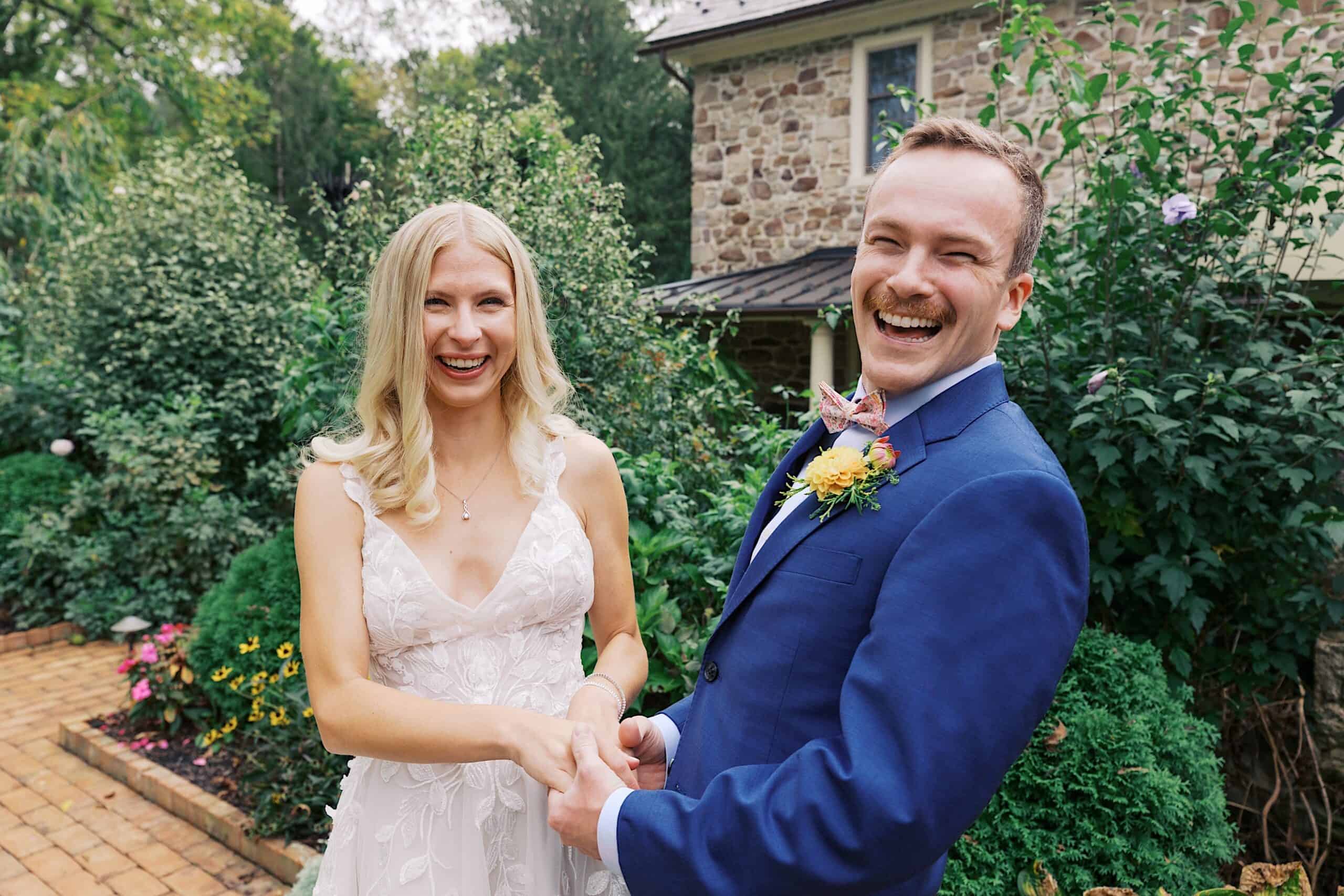 A bride in a white dress and a groom in a blue suit stand smiling and holding hands outdoors, celebrating their beautiful wedding at Vincent Forge Mansion, with a stone house in the background.