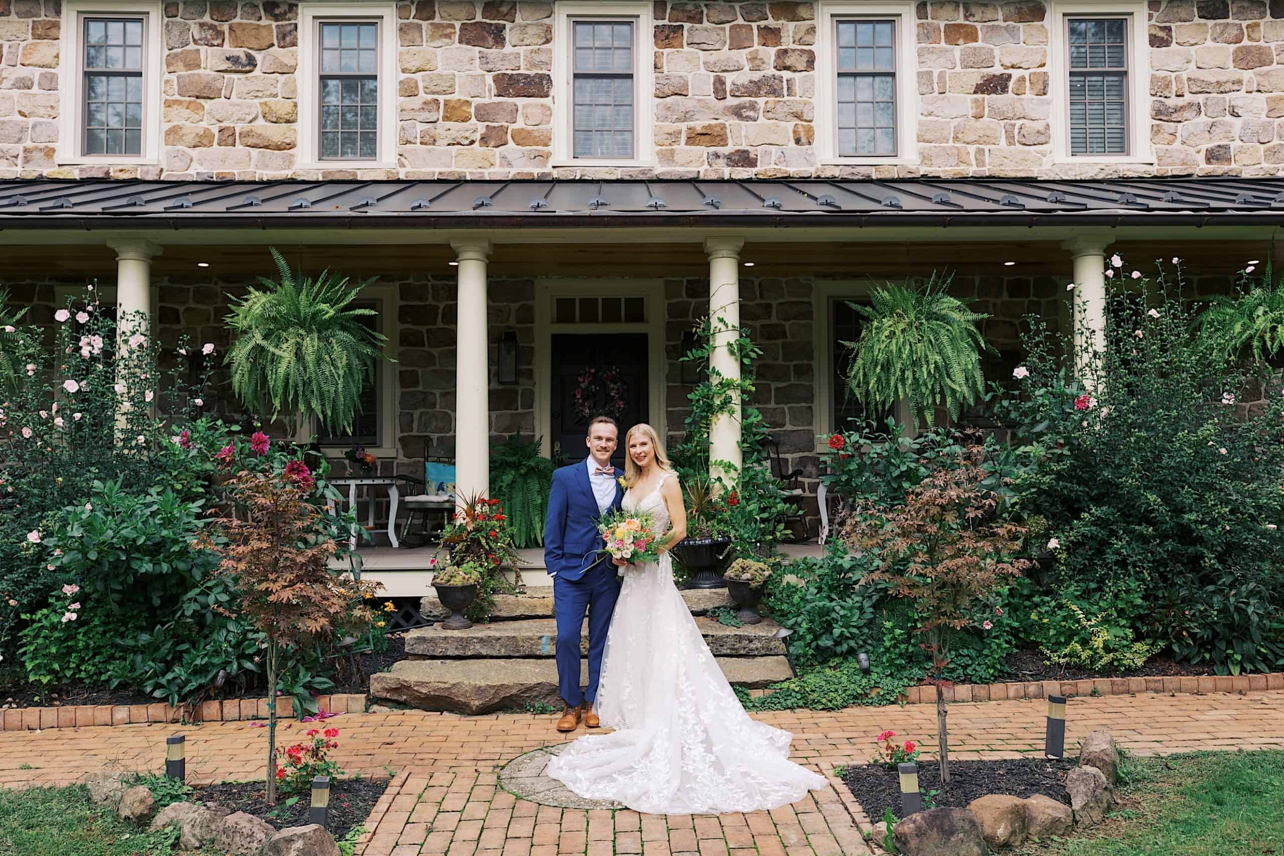 A bride and groom stand together on a brick pathway in front of a stone house with a covered porch and lush garden landscaping, celebrating their beautiful wedding at Vincent Forge Mansion.