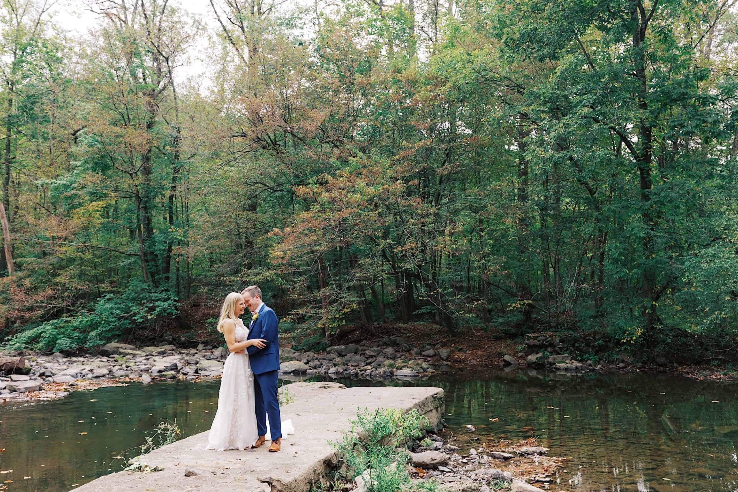 A couple stands embracing on a concrete platform over a shallow stream, surrounded by dense green trees—capturing the romance of a beautiful wedding at Vincent Forge Mansion in this enchanting forest setting.