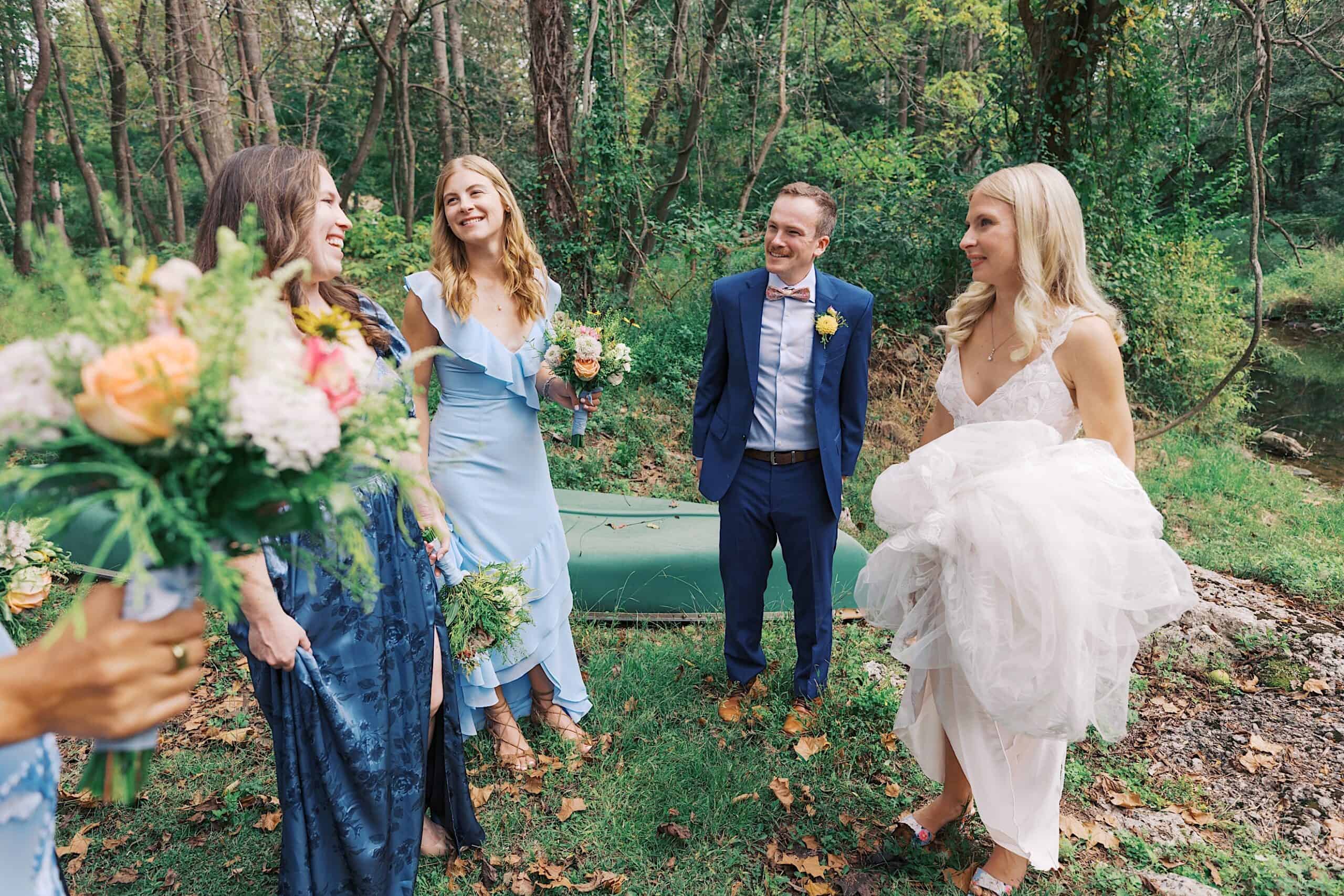 Four people dressed in formal attire stand outdoors near a canoe, holding bouquets and smiling, celebrating a beautiful wedding at Vincent Forge Mansion, with trees and a stream in the background.