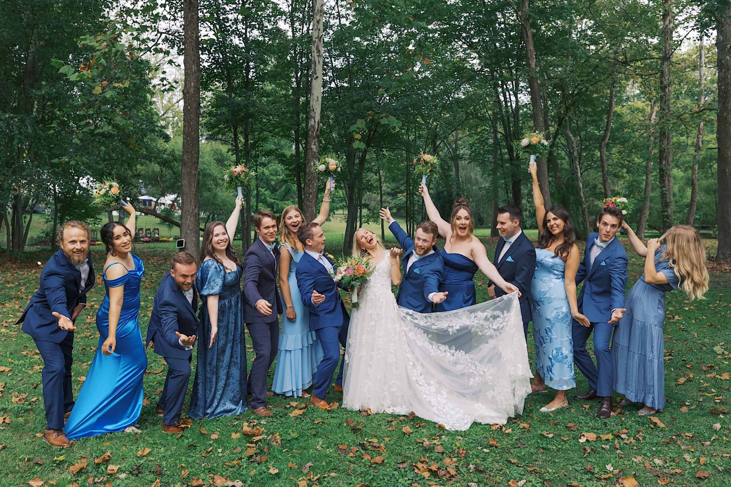 A wedding party poses outdoors in a wooded area at the beautiful wedding at Vincent Forge Mansion, with the bride and groom at the center surrounded by bridesmaids in blue dresses and groomsmen in blue suits.