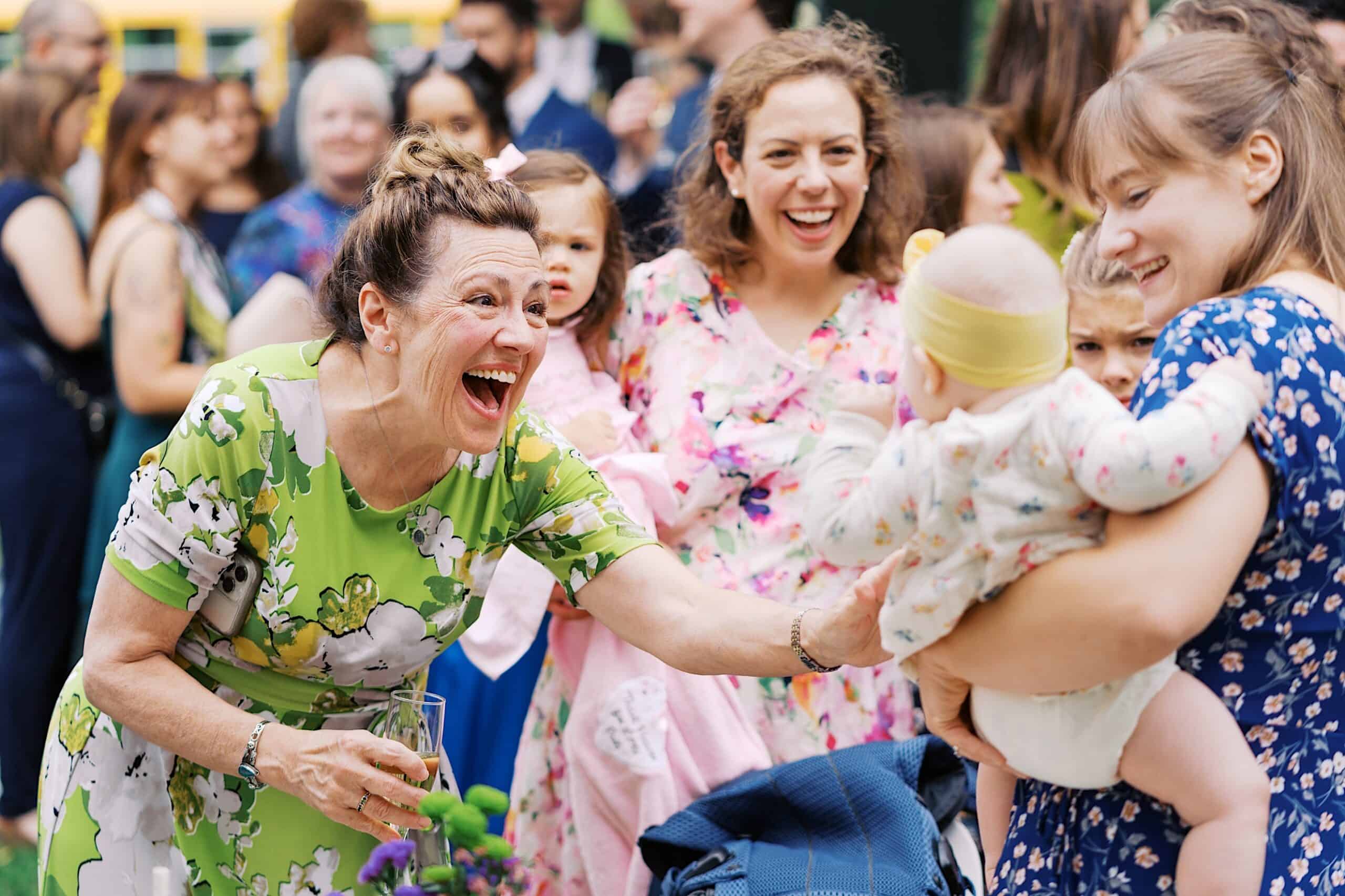 At a beautiful wedding at Vincent Forge Mansion, a woman in a green floral dress smiles and reaches toward a baby held by another woman in a blue dress, surrounded by a joyous crowd at the celebration.