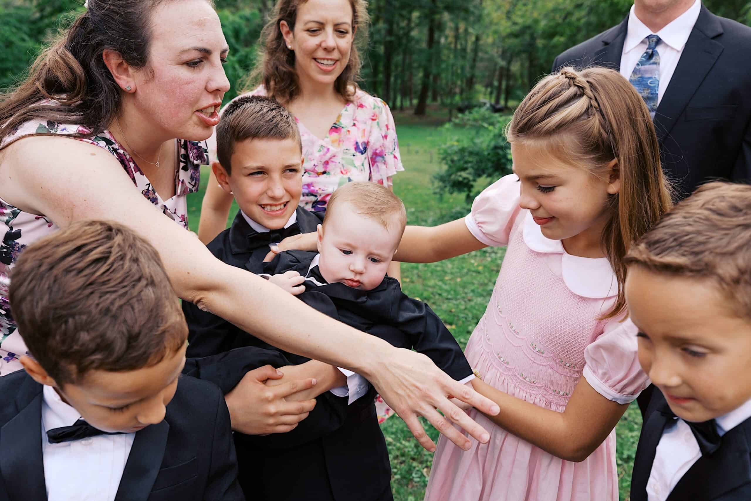 A group of children and adults, dressed formally, gather outside at a beautiful wedding at Vincent Forge Mansion. One woman steadies a baby in a suit as other children and a man in a suit interact with them. Trees are in the background.