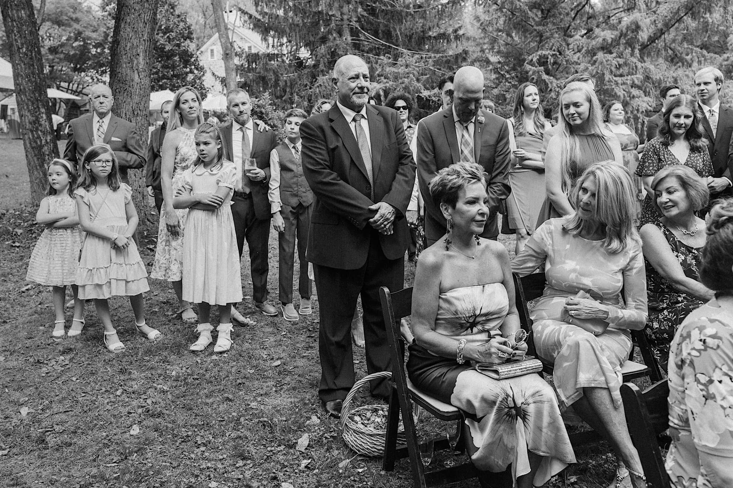 A group of people, both seated and standing, gather outdoors among trees in formal attire, attending a beautiful wedding at Vincent Forge Mansion.