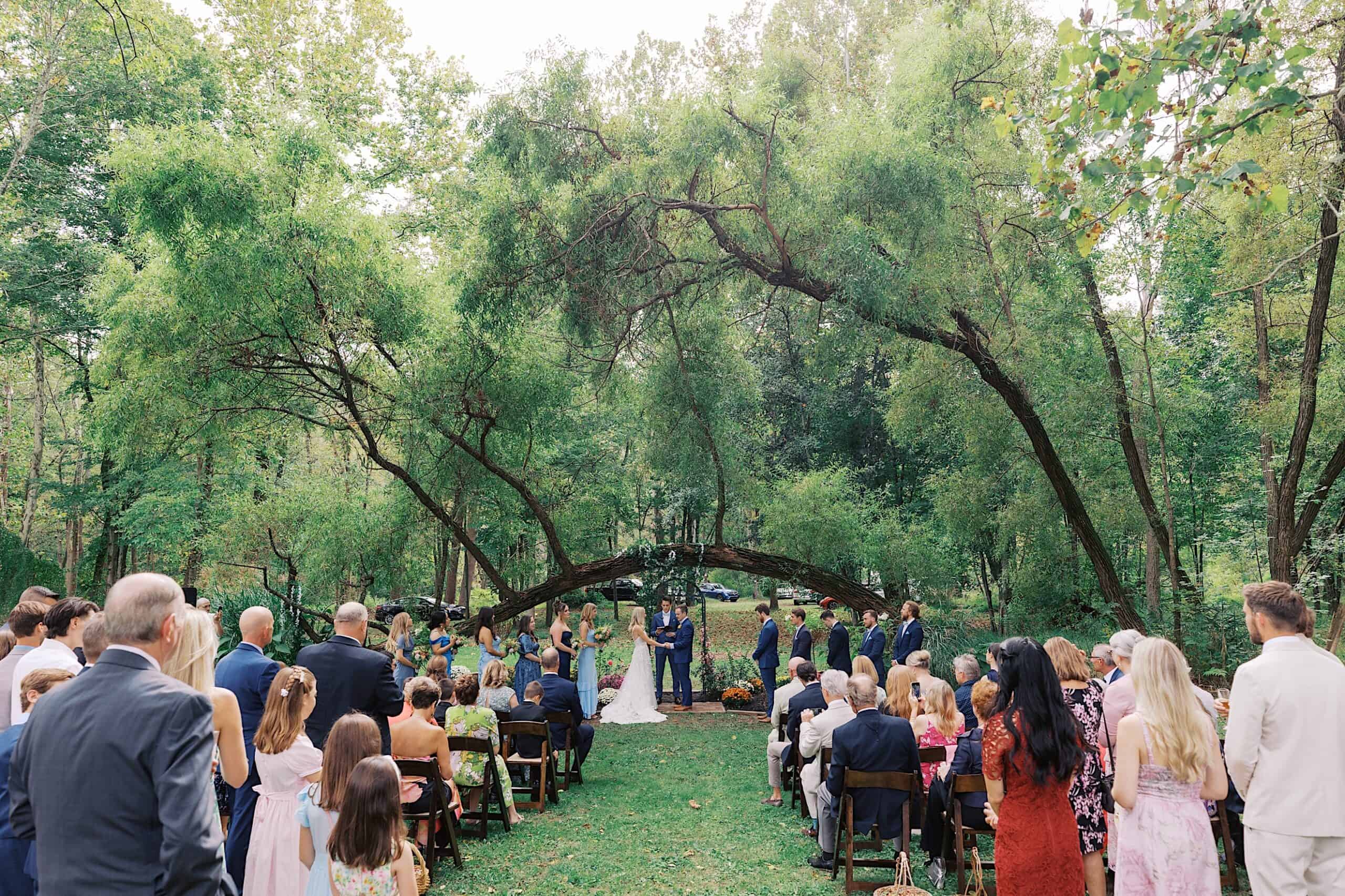 A beautiful wedding at Vincent Forge Mansion is taking place outdoors under large trees, with guests seated on either side and the couple standing at the front with their officiant.