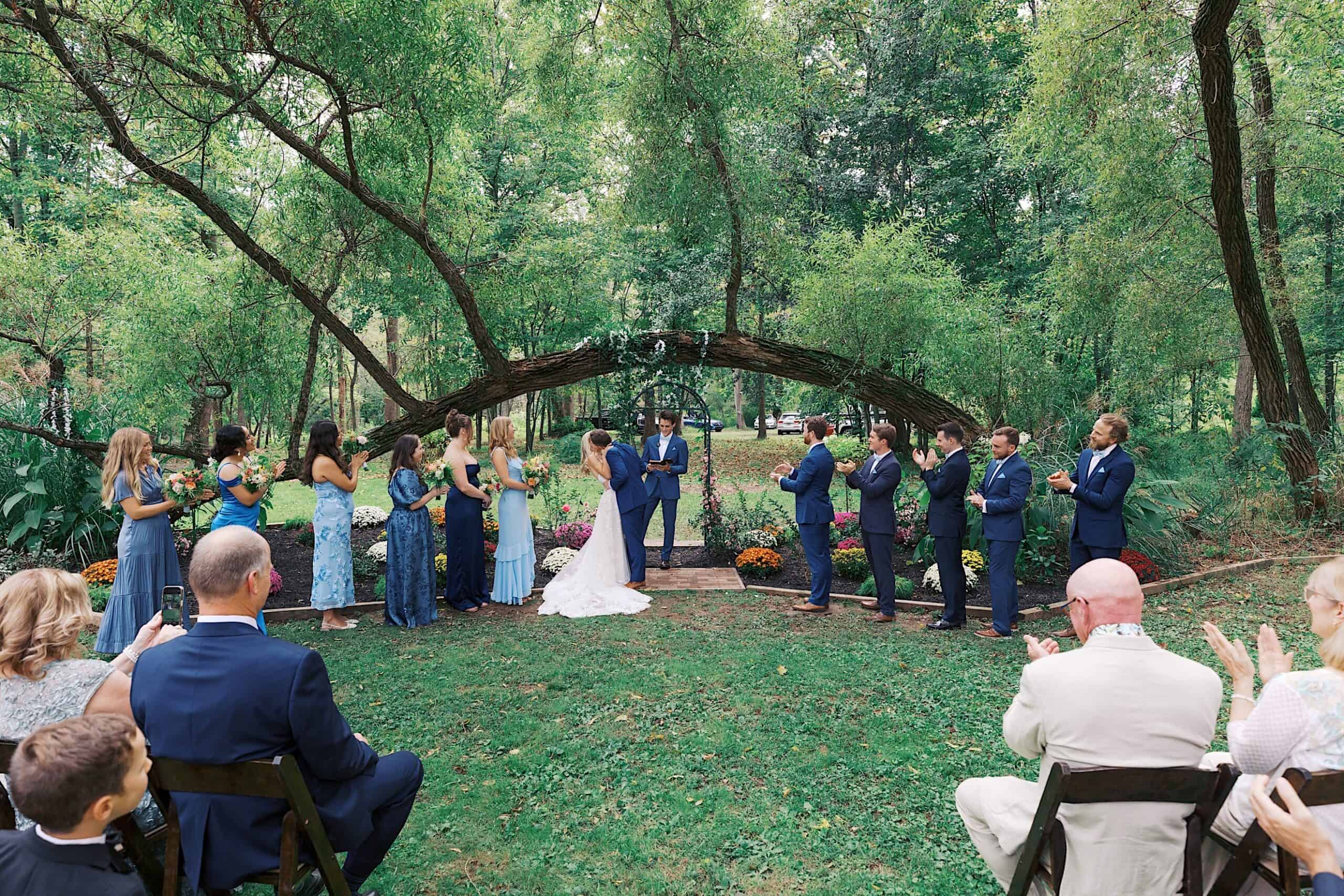 A bride and groom stand at an outdoor altar surrounded by trees, exchanging vows during a beautiful wedding at Vincent Forge Mansion, with their wedding party and seated guests present.