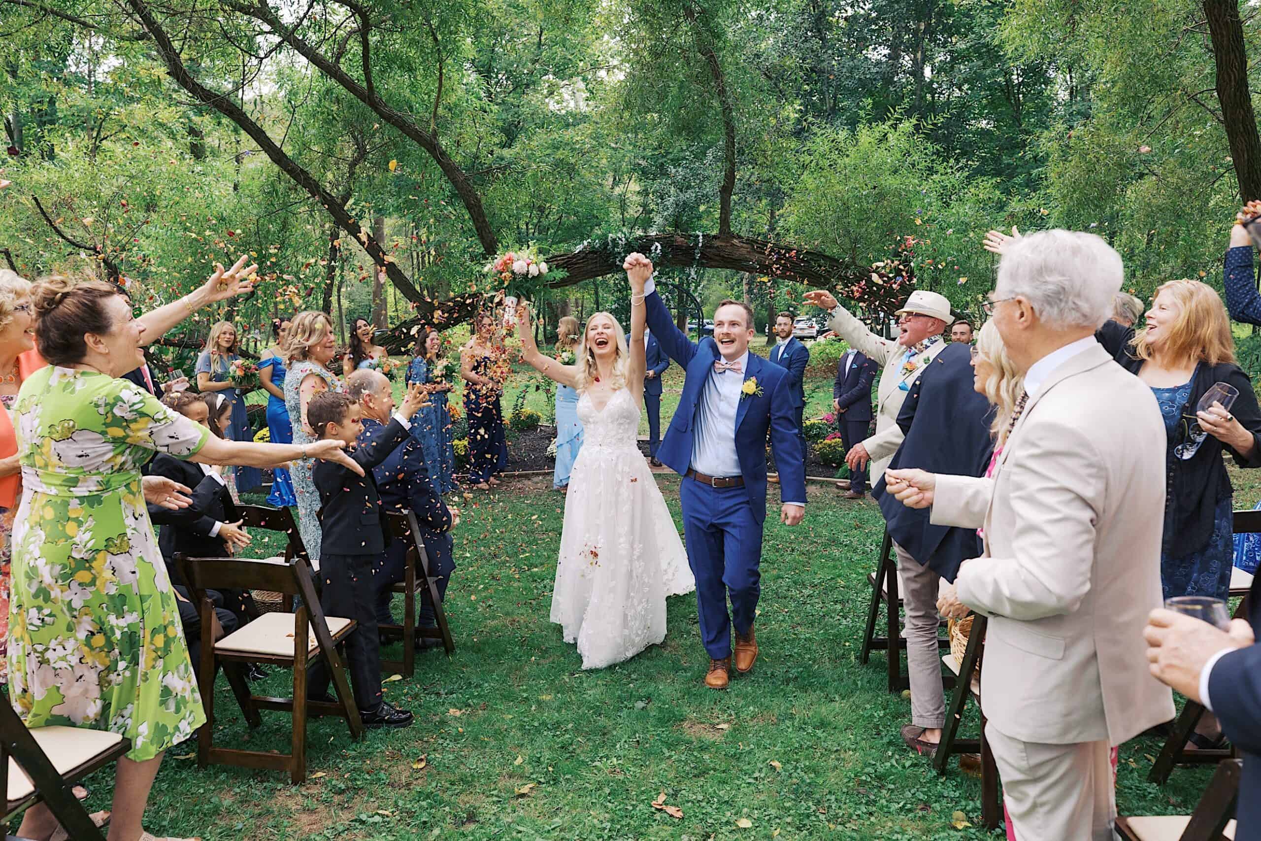 A bride and groom walk down an outdoor aisle at the beautiful wedding at Vincent Forge Mansion as guests throw flower petals and clap, surrounded by trees and greenery.