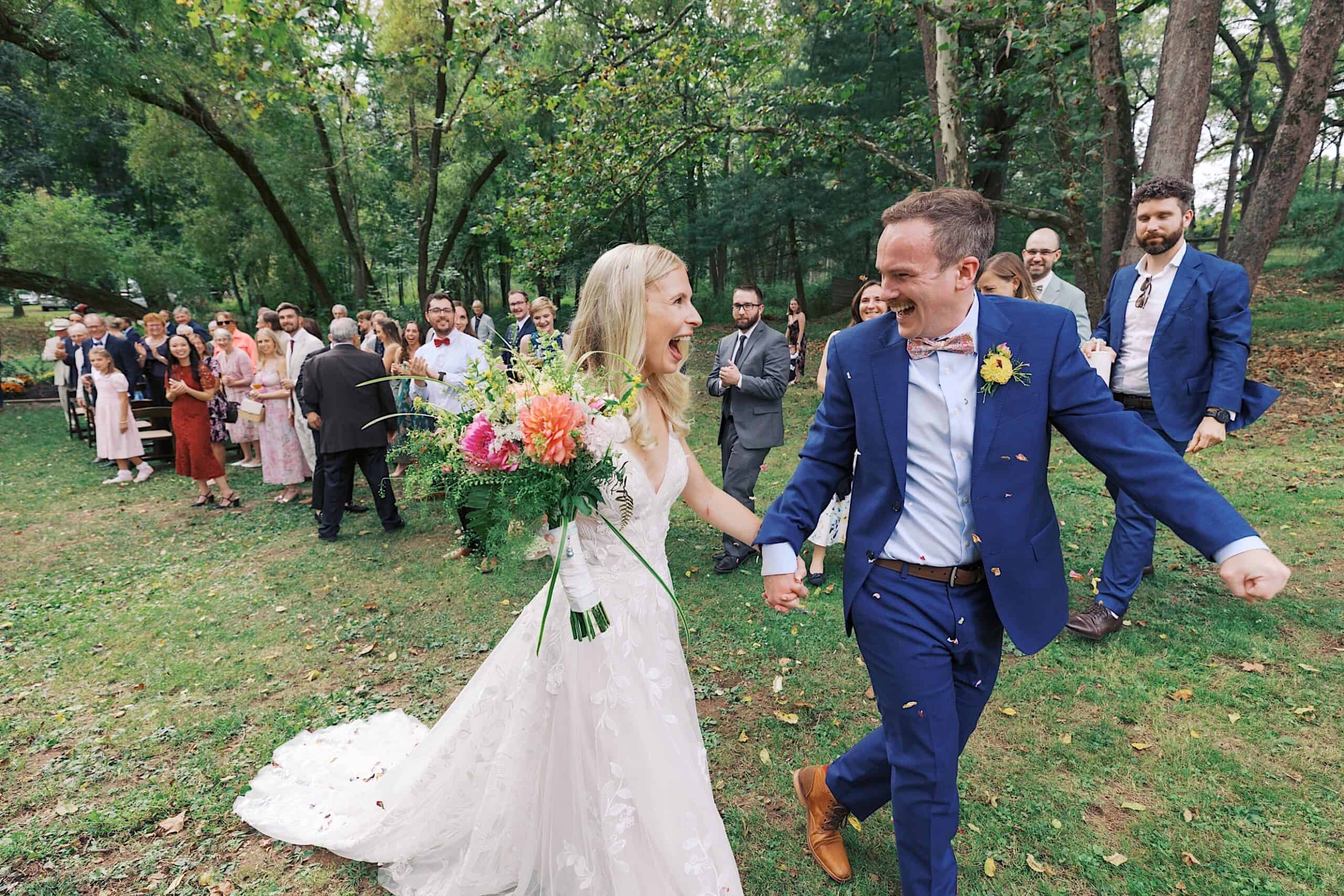 A bride and groom holding hands walk through a grassy outdoor area at their beautiful wedding at Vincent Forge Mansion, smiling, as a group of guests stands behind them clapping.
