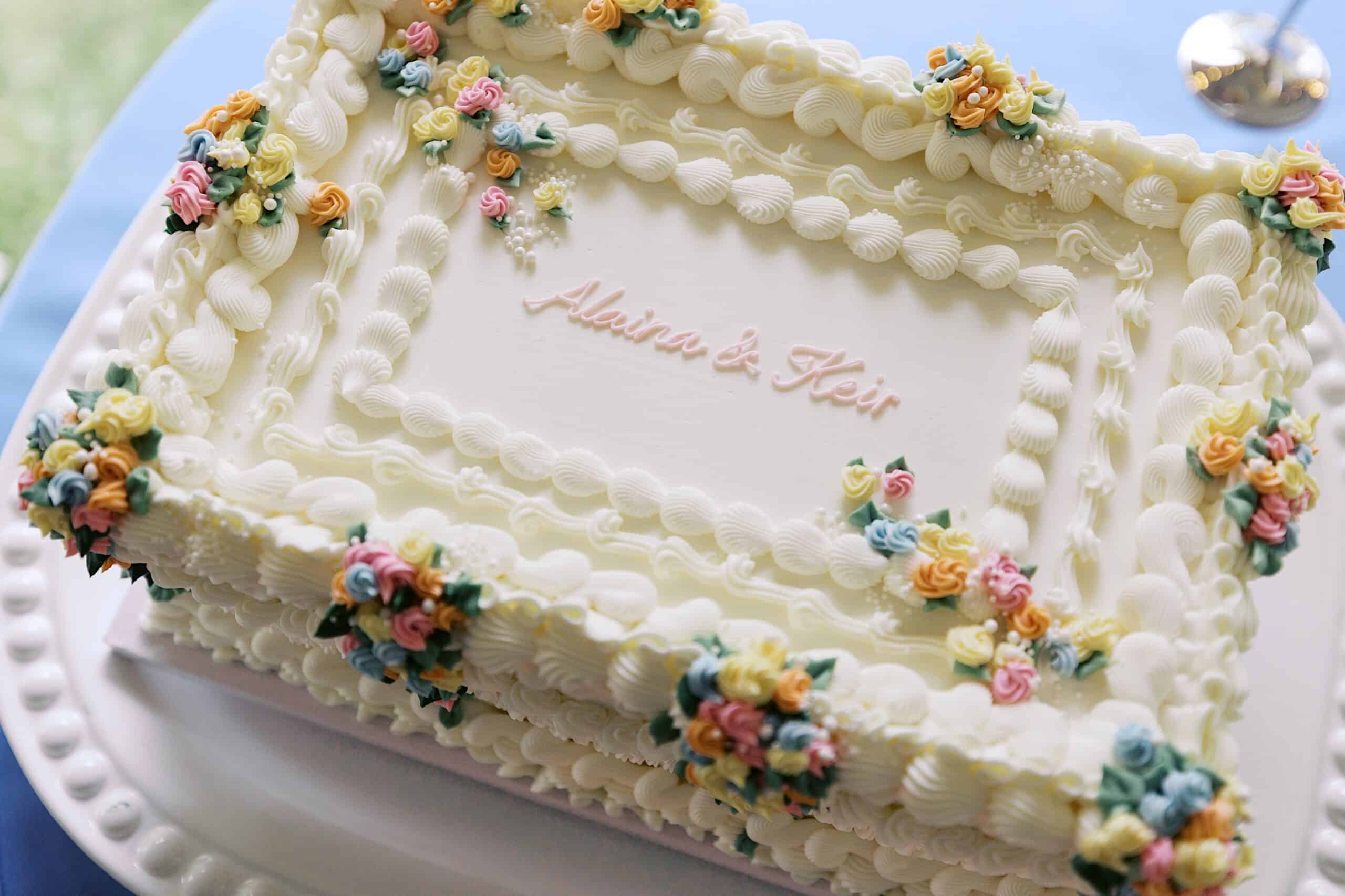 A rectangular white cake decorated with pastel flowers and piped borders, featuring the names "Adaira & Keir" written in pink icing on top—perfect for a beautiful wedding at Vincent Forge Mansion.
