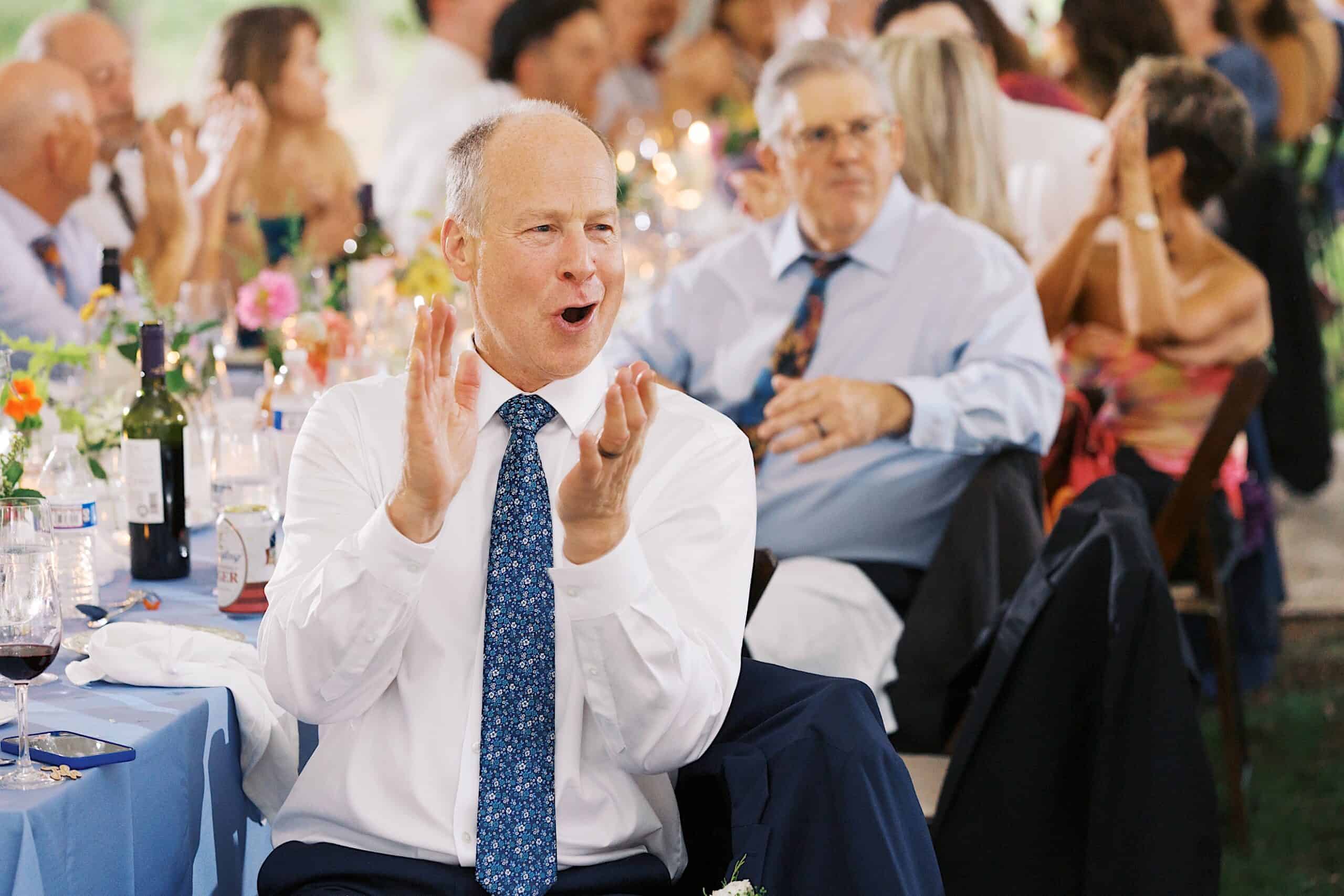 A man in a white shirt and blue tie claps while seated at a table during a beautiful wedding at Vincent Forge Mansion, surrounded by other guests.