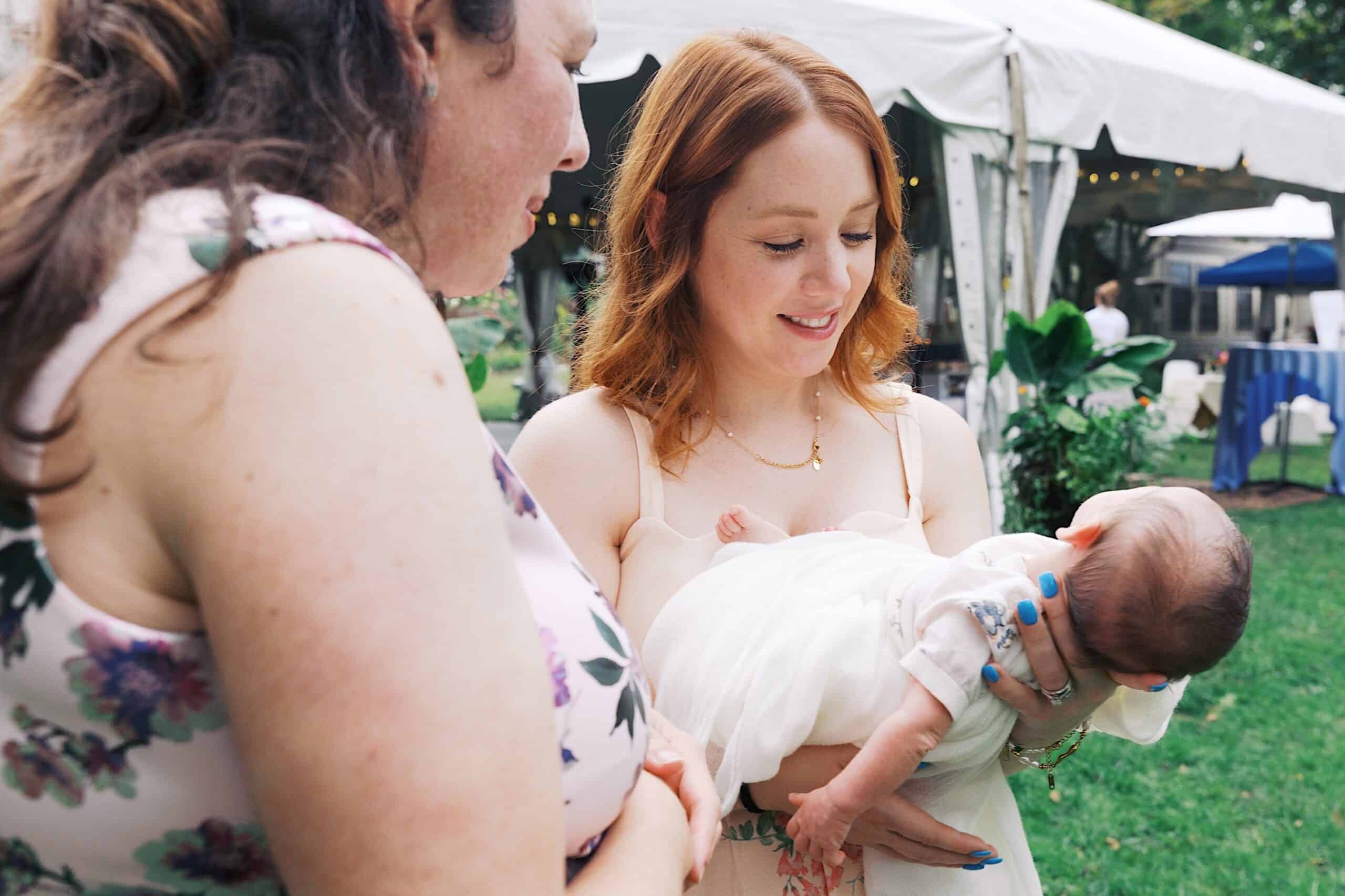 Two women stand outdoors at a beautiful wedding at Vincent Forge Mansion; one holds a baby in a white outfit while the other looks on. A white tent and lush greenery provide a charming backdrop.